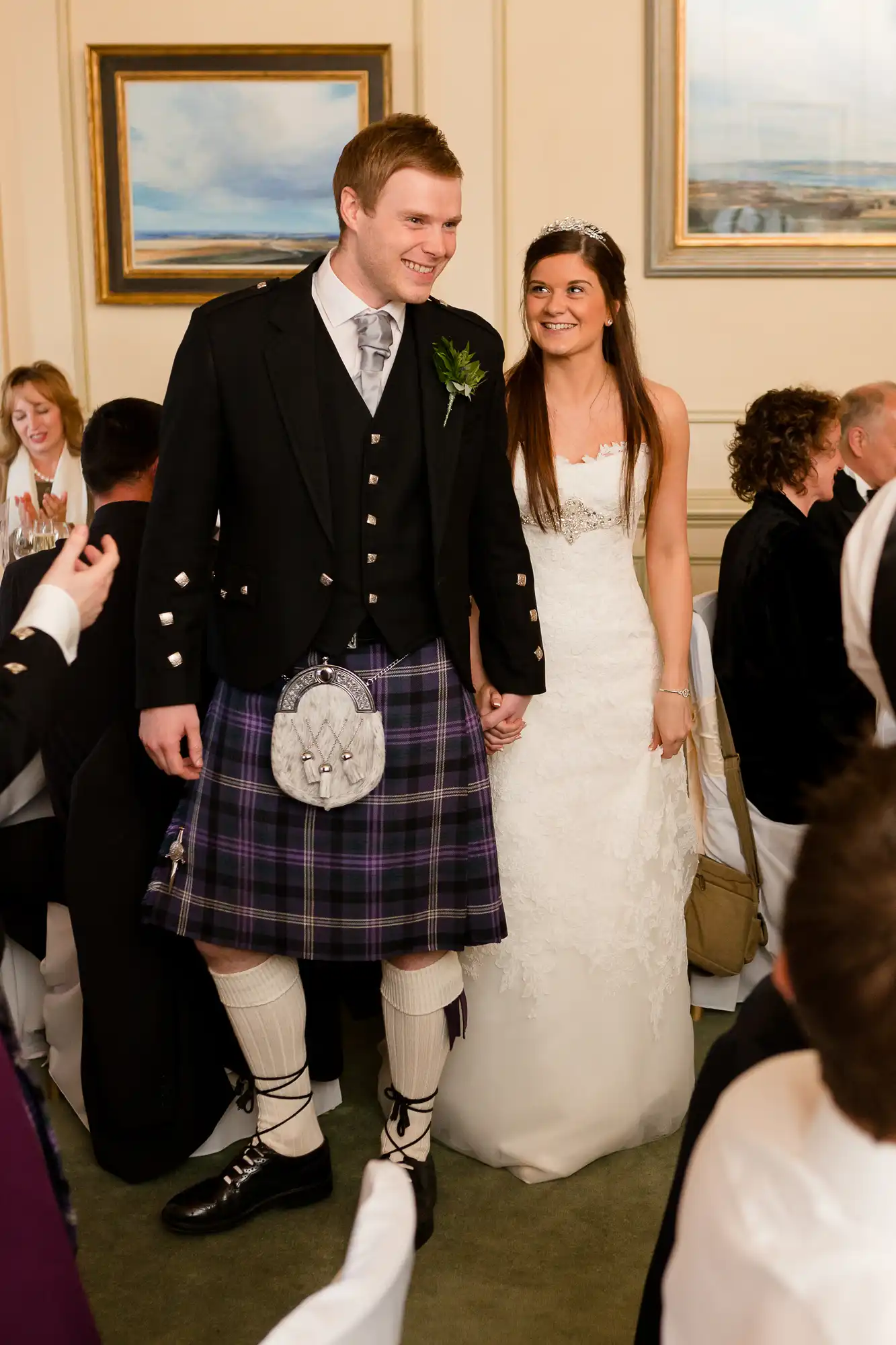 A bride in a white wedding dress and a groom in a traditional scottish kilt and jacket smile as they walk through a room of seated guests.