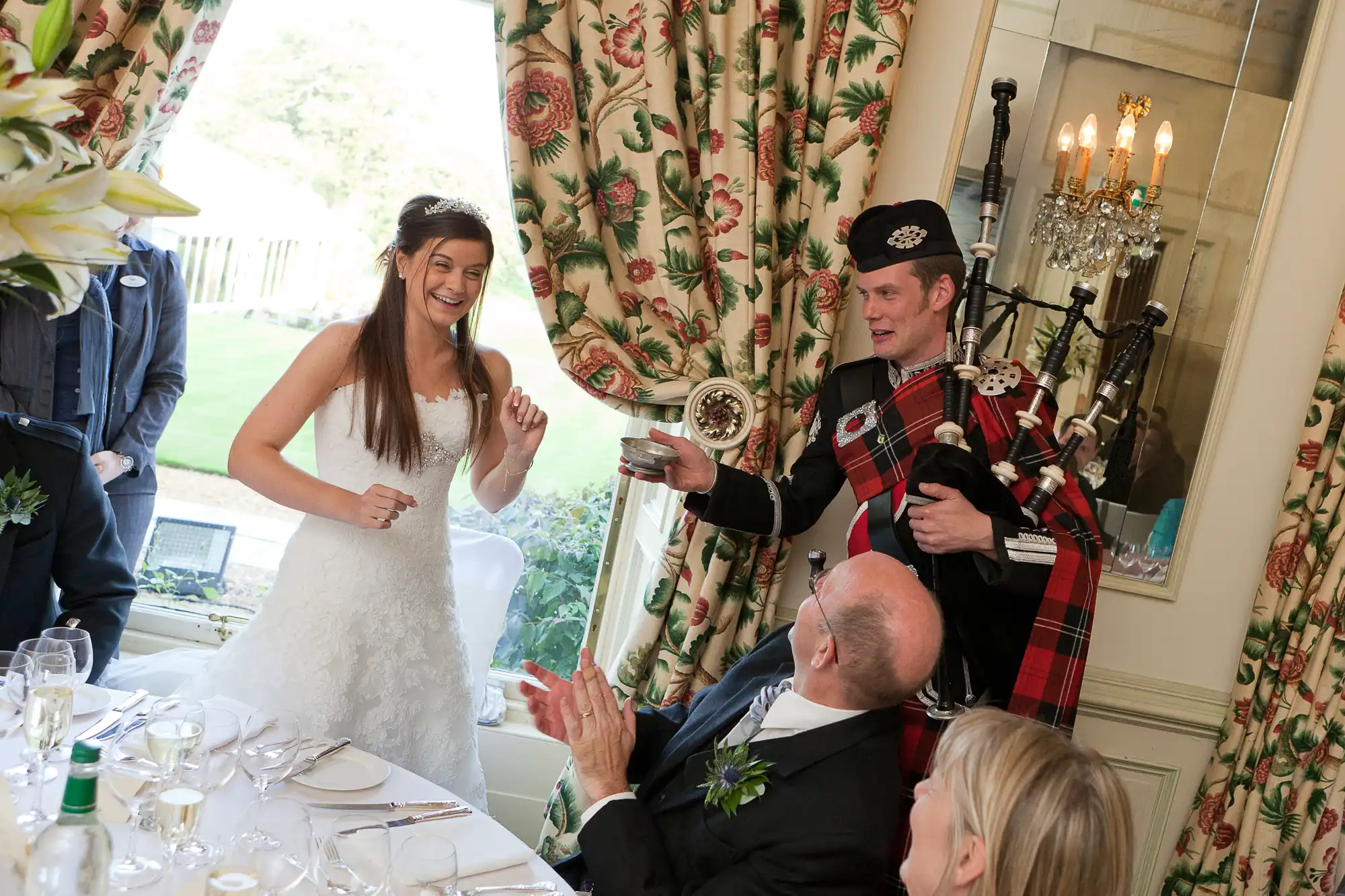 A bride laughs joyfully beside a bagpiper in a kilt at a wedding reception, with guests and elegant decor around them.