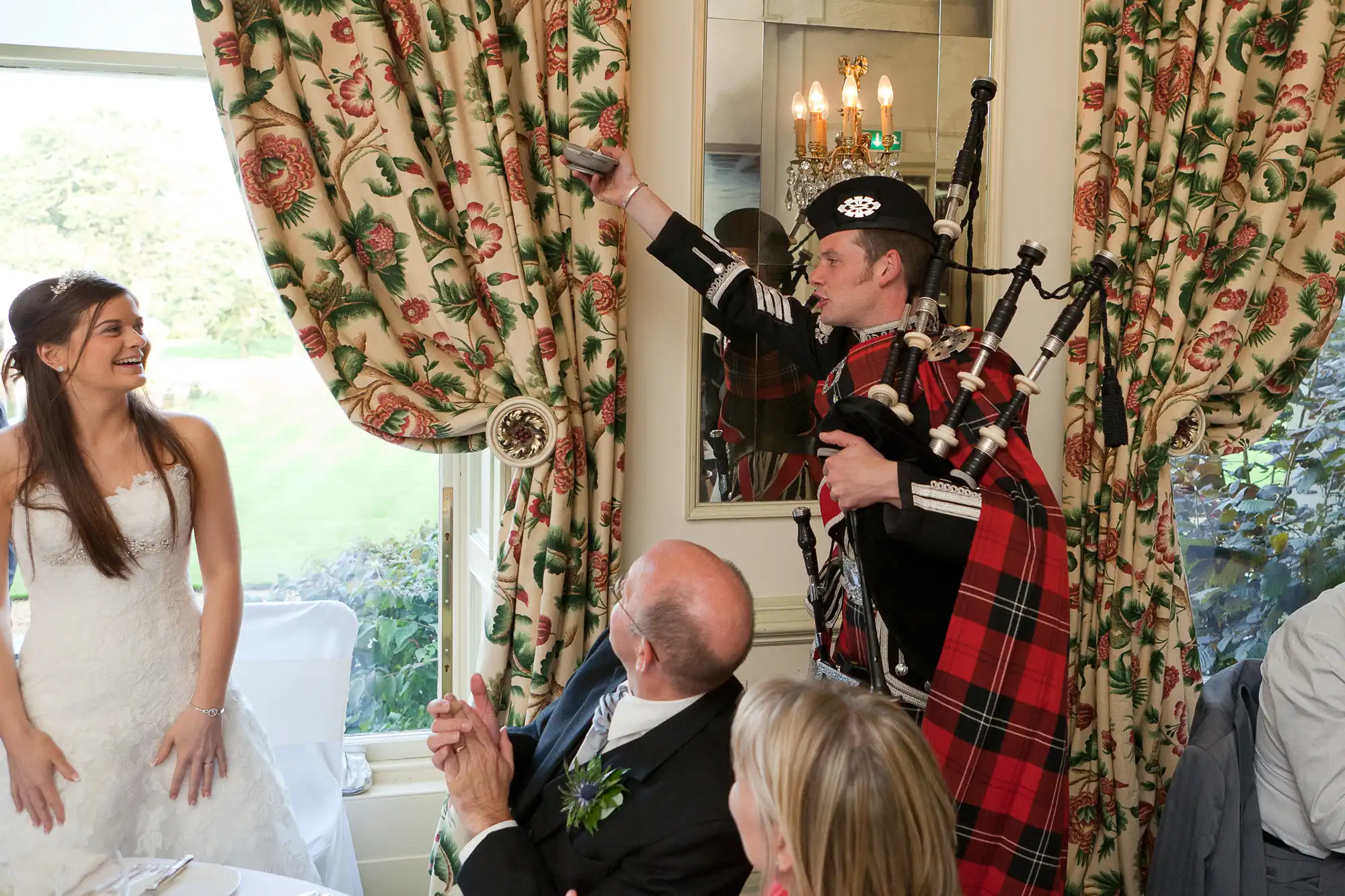 A bride laughs as a bagpiper in traditional scottish attire plays indoors by a window during a wedding reception.