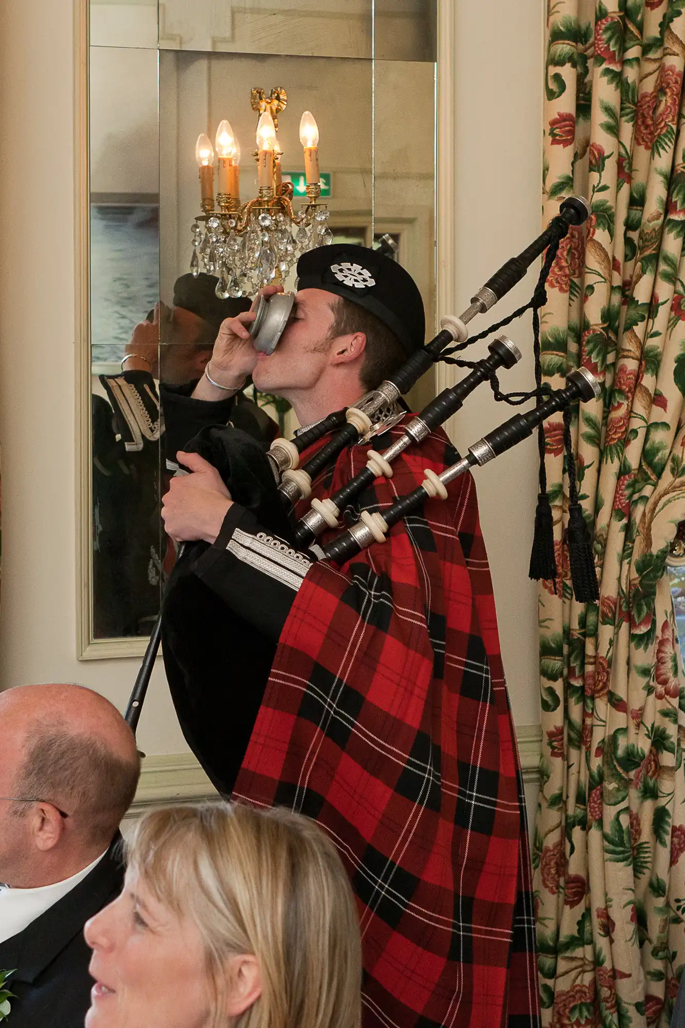 A man in traditional scottish attire, including a kilt and tartan plaid, drinks from a cup while holding bagpipes at a formal indoor event.