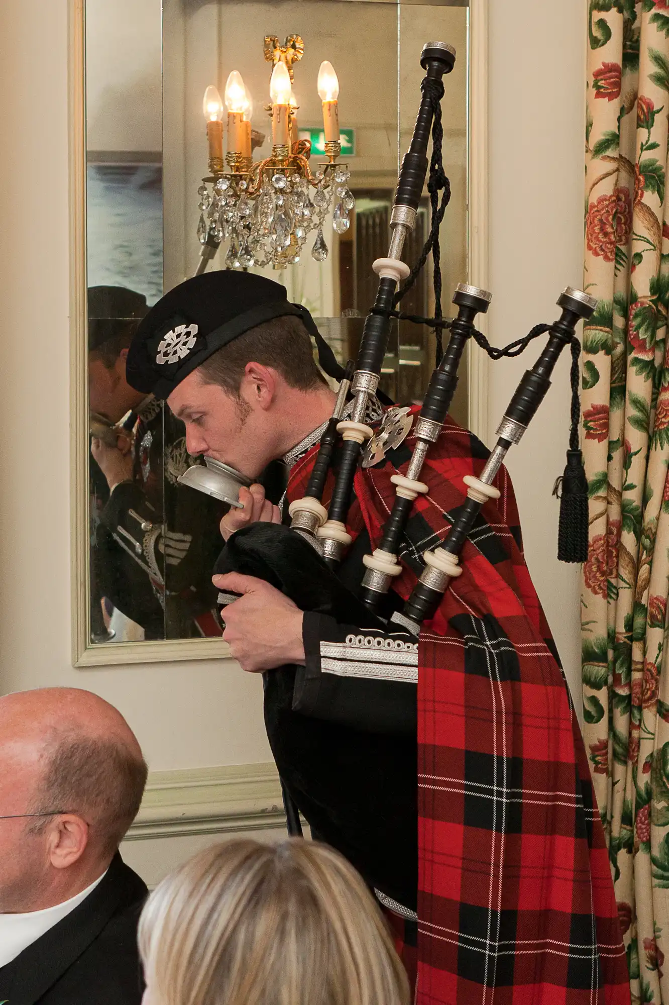 A man in traditional scottish attire plays the bagpipes indoors at a formal event, standing next to seated guests.