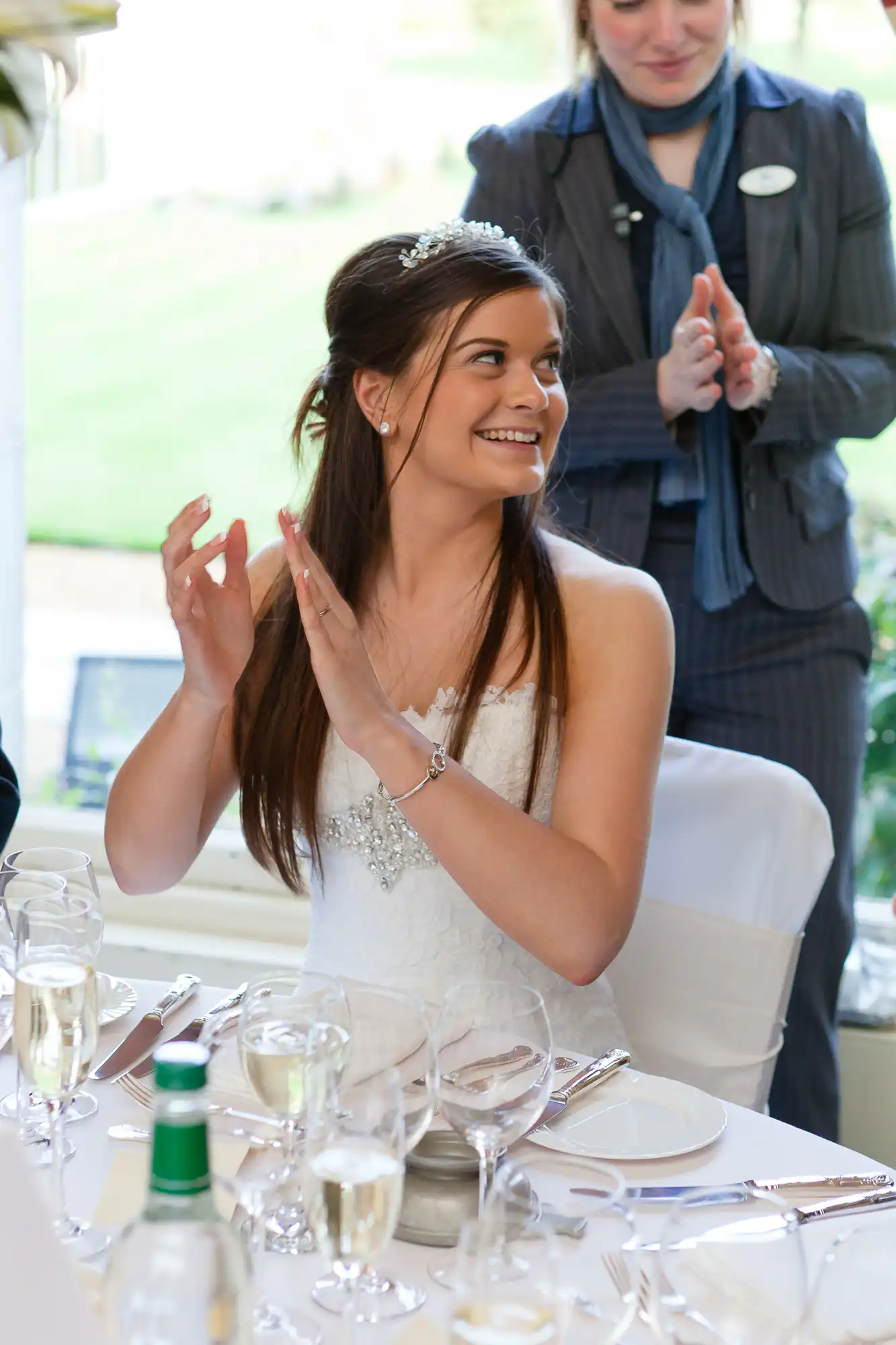 A bride in a white dress, seated and clapping at a table during a wedding reception, with a smiling expression and a server in the background.