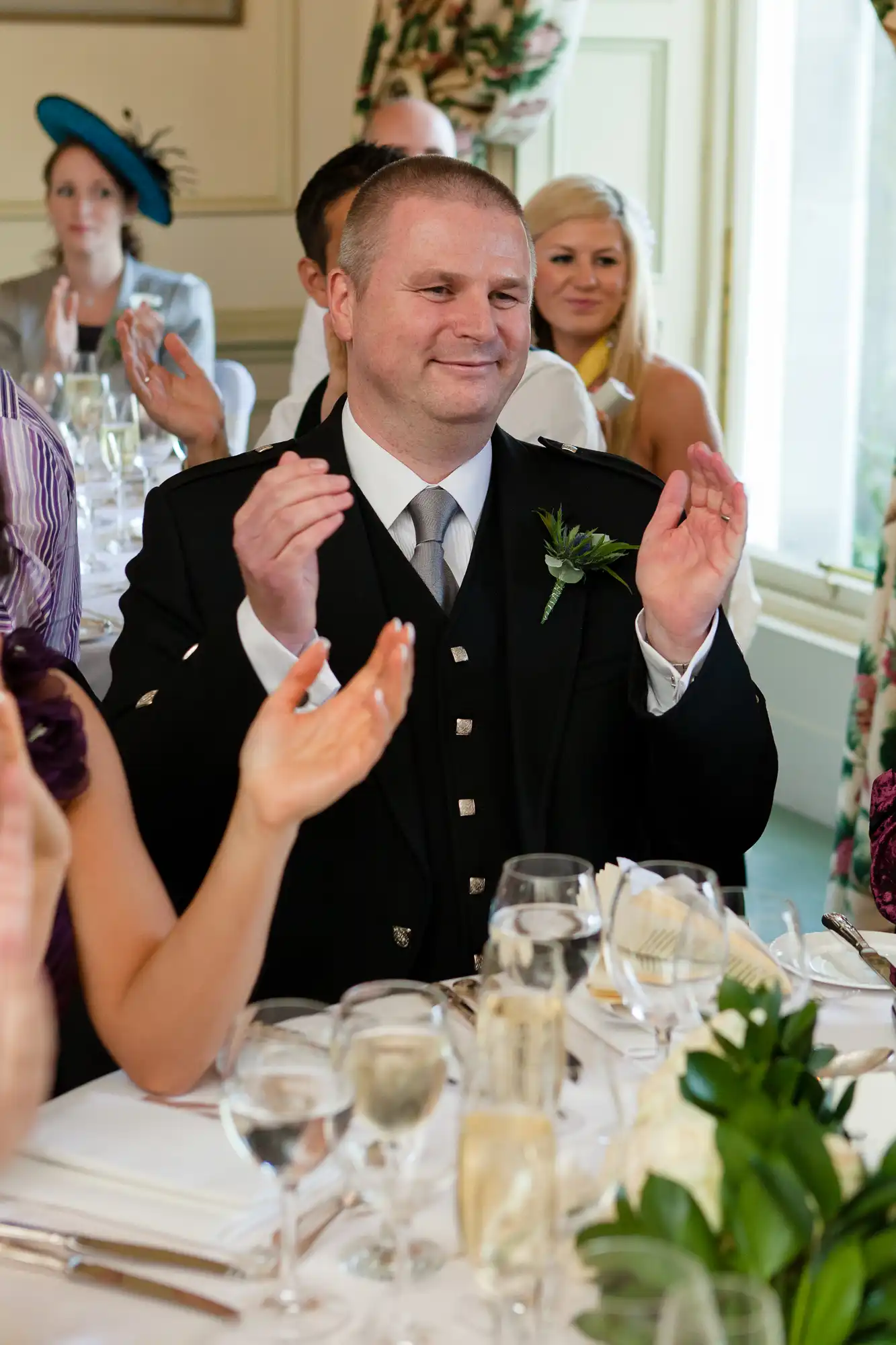 Man in formal attire with a boutonniere, clapping at a dining table during an event, with guests in the background.
