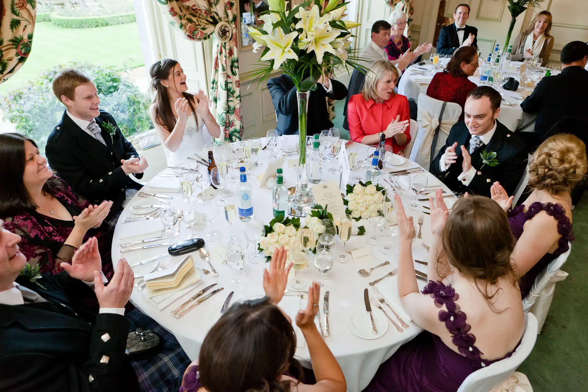 Guests at a wedding reception seated around a large table adorned with flowers, clapping and interacting joyfully.