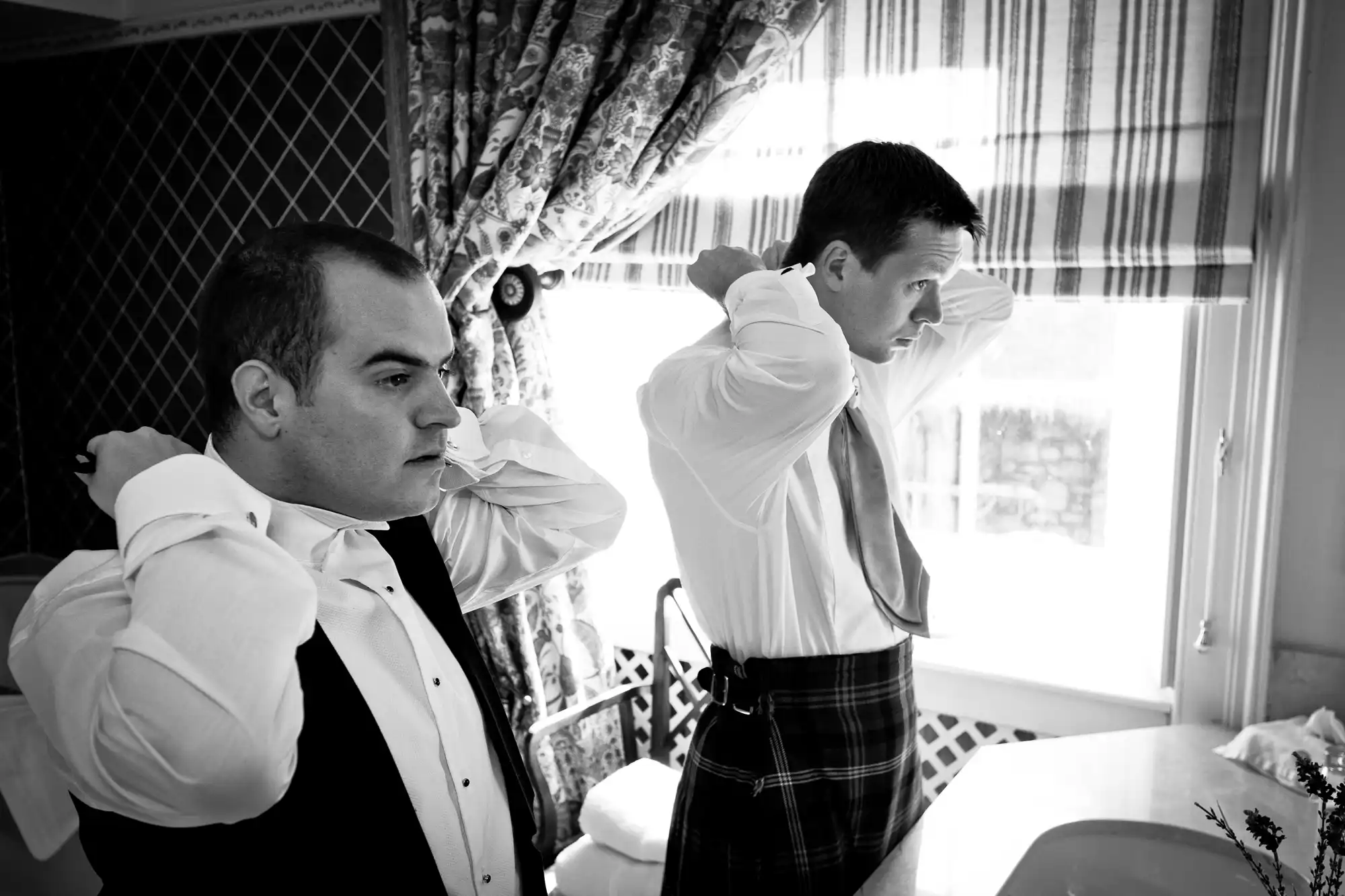 Two men in formal attire adjusting their bow ties in a room with floral curtains, one wearing a kilt.