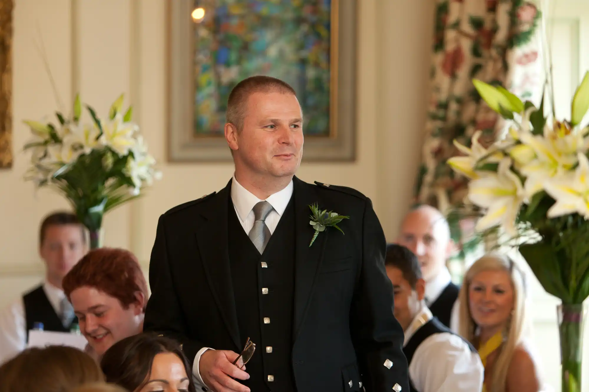 A man in a formal black suit with a boutonniere stands in a room with seated guests and white lilies, looking proud and emotional.