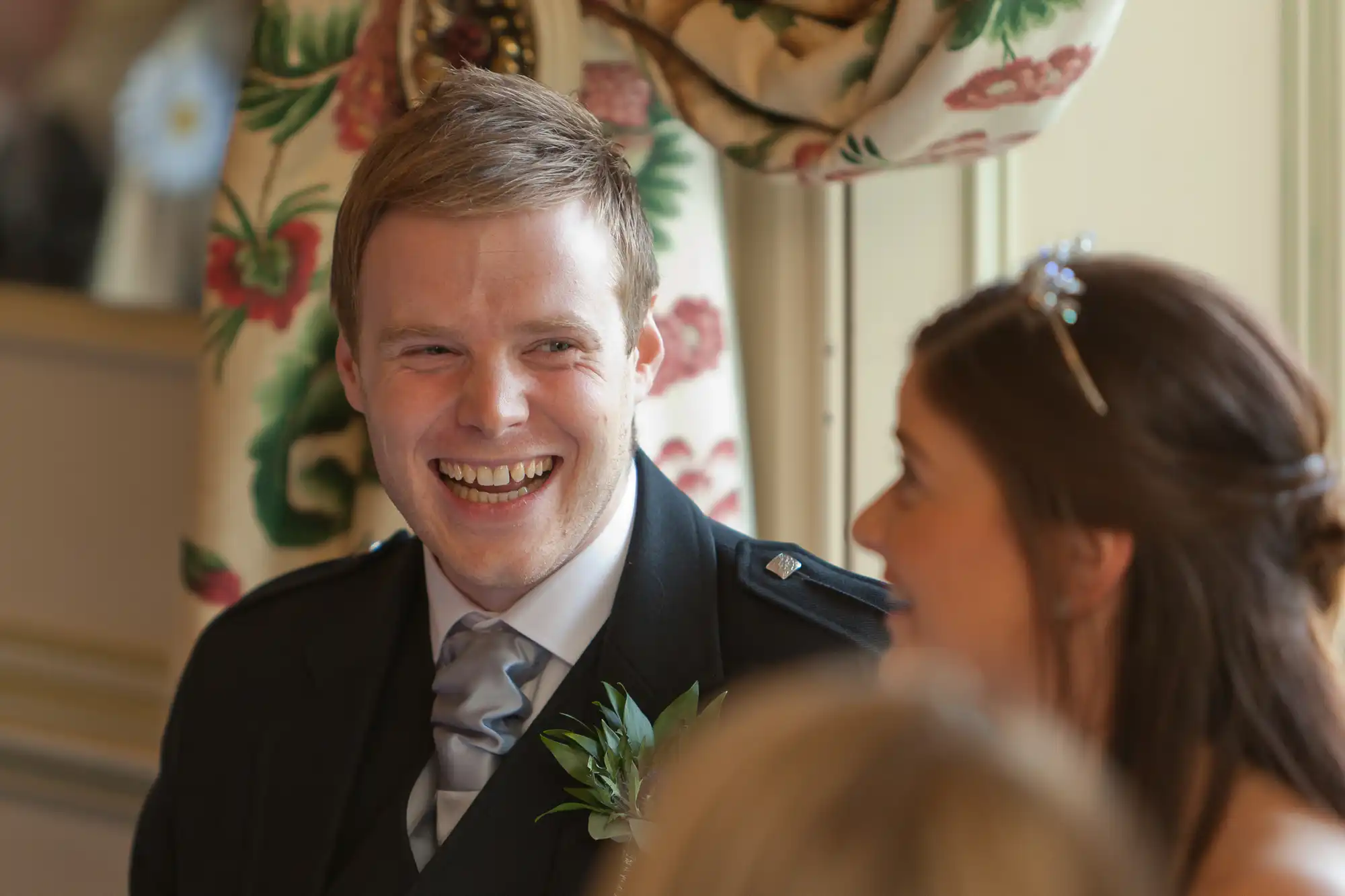 A man in a suit and tie, smiling broadly at a woman with a tiara, visible in partial profile, in a room with floral wallpaper.