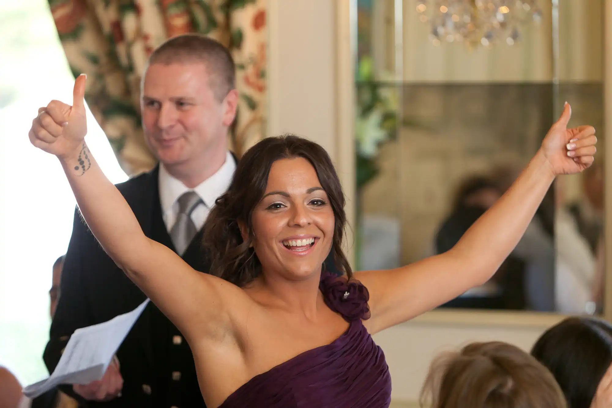 Woman in a purple dress giving thumbs up with a smiling man in the background at a formal event.