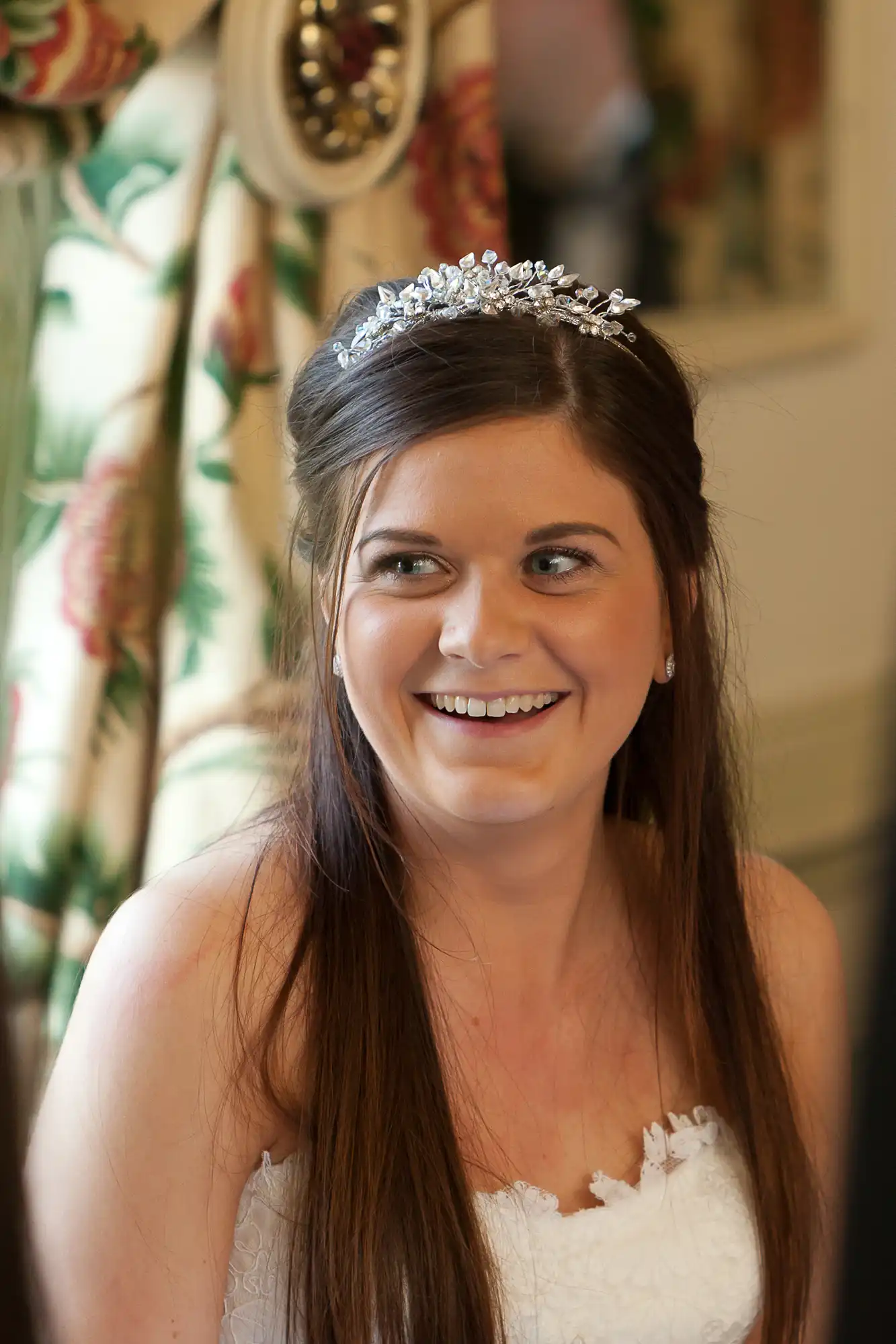A smiling bride wearing a tiara sits in a room, with white floral dress details visible. .
