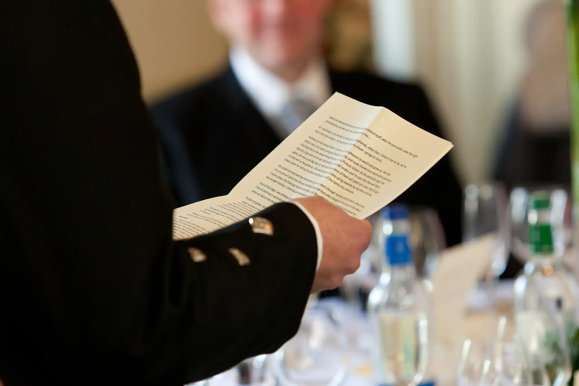 A person holding a document at a formal event, with blurred individuals in the background. focus is on the text of the document.