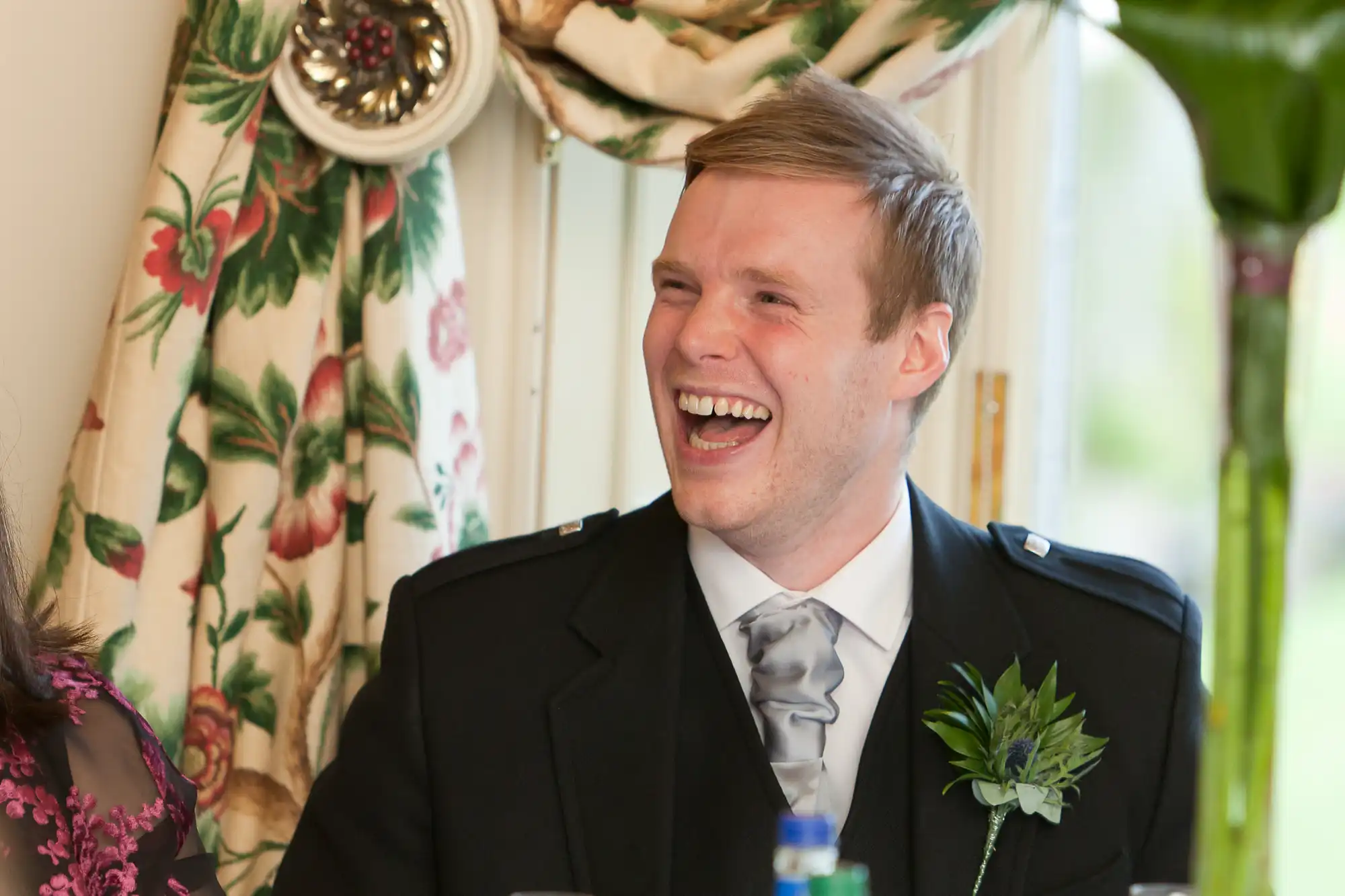 A man in a formal suit and tie laughing joyously at a table decorated with a floral curtain backdrop.