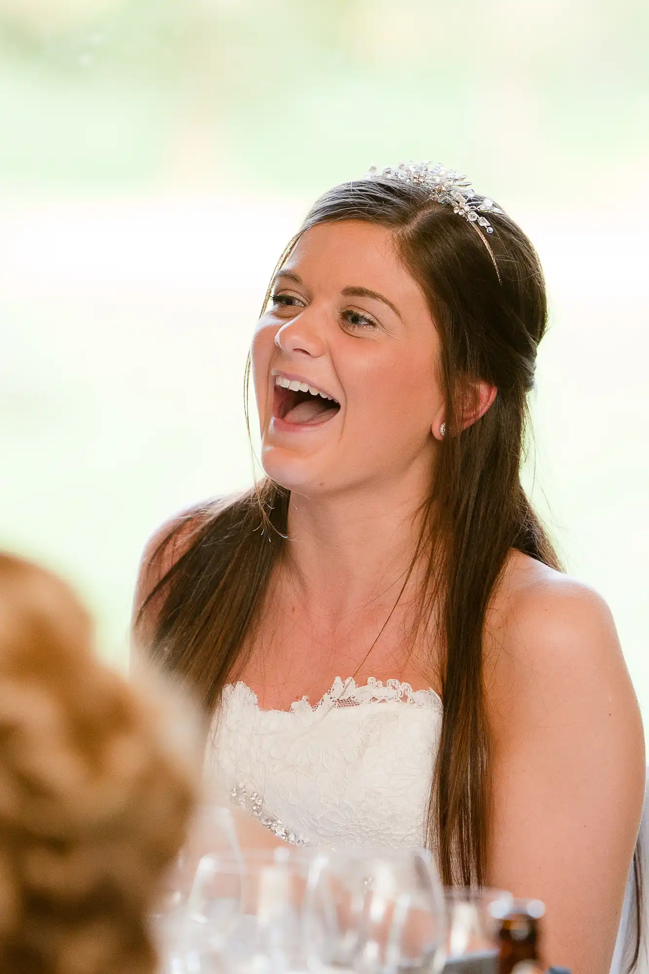 A joyful bride in a white dress with a tiara laughing at a wedding reception.