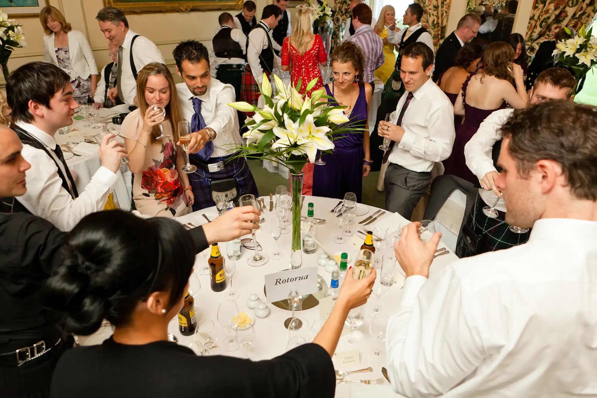 Group of people engaging in conversation and toasting drinks at a festive table with floral centerpieces in an elegant room.