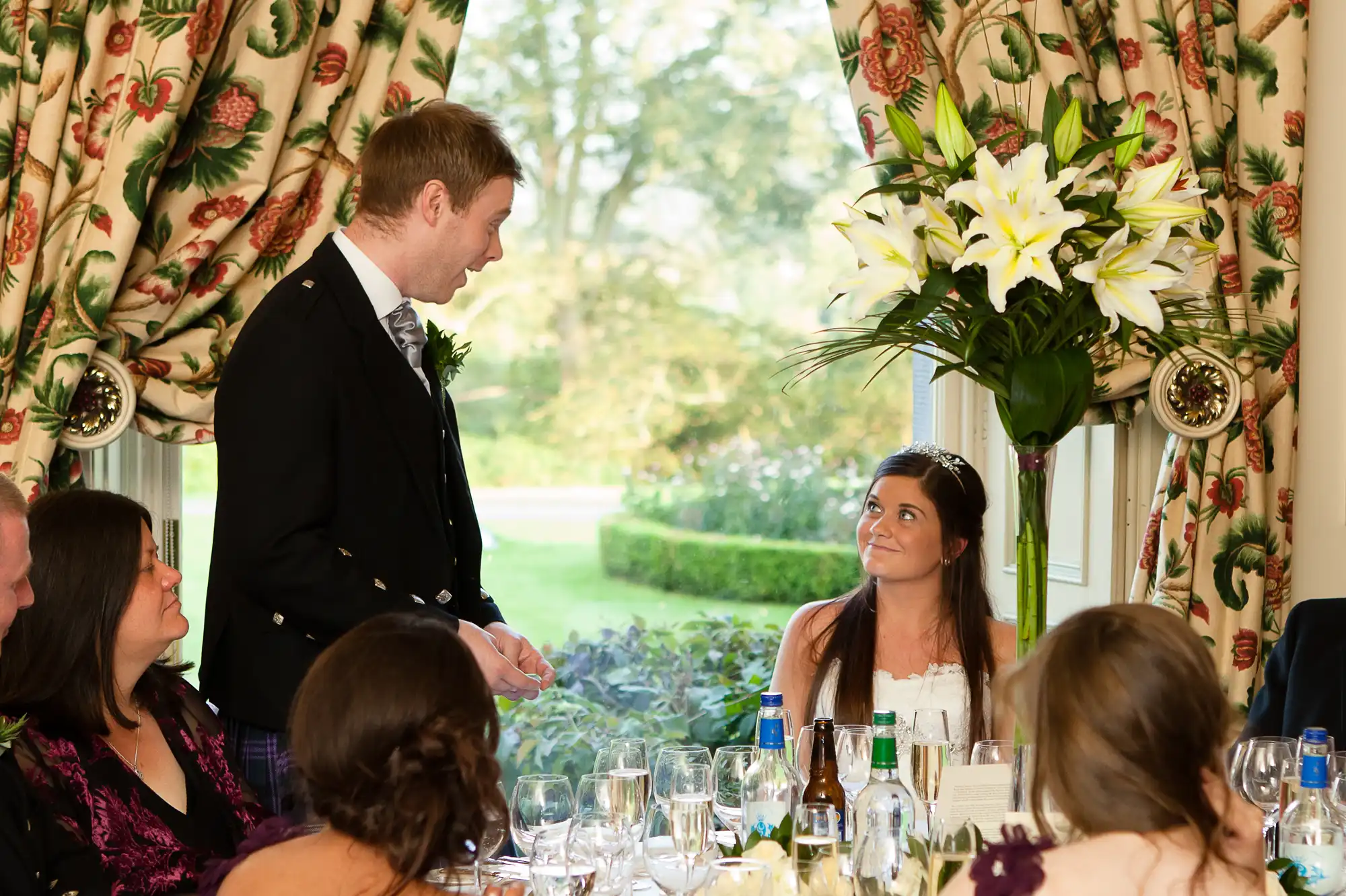 A man in a suit speaks to a woman in a white dress and headband at a wedding reception, with guests around a table and large lilies in the background.
