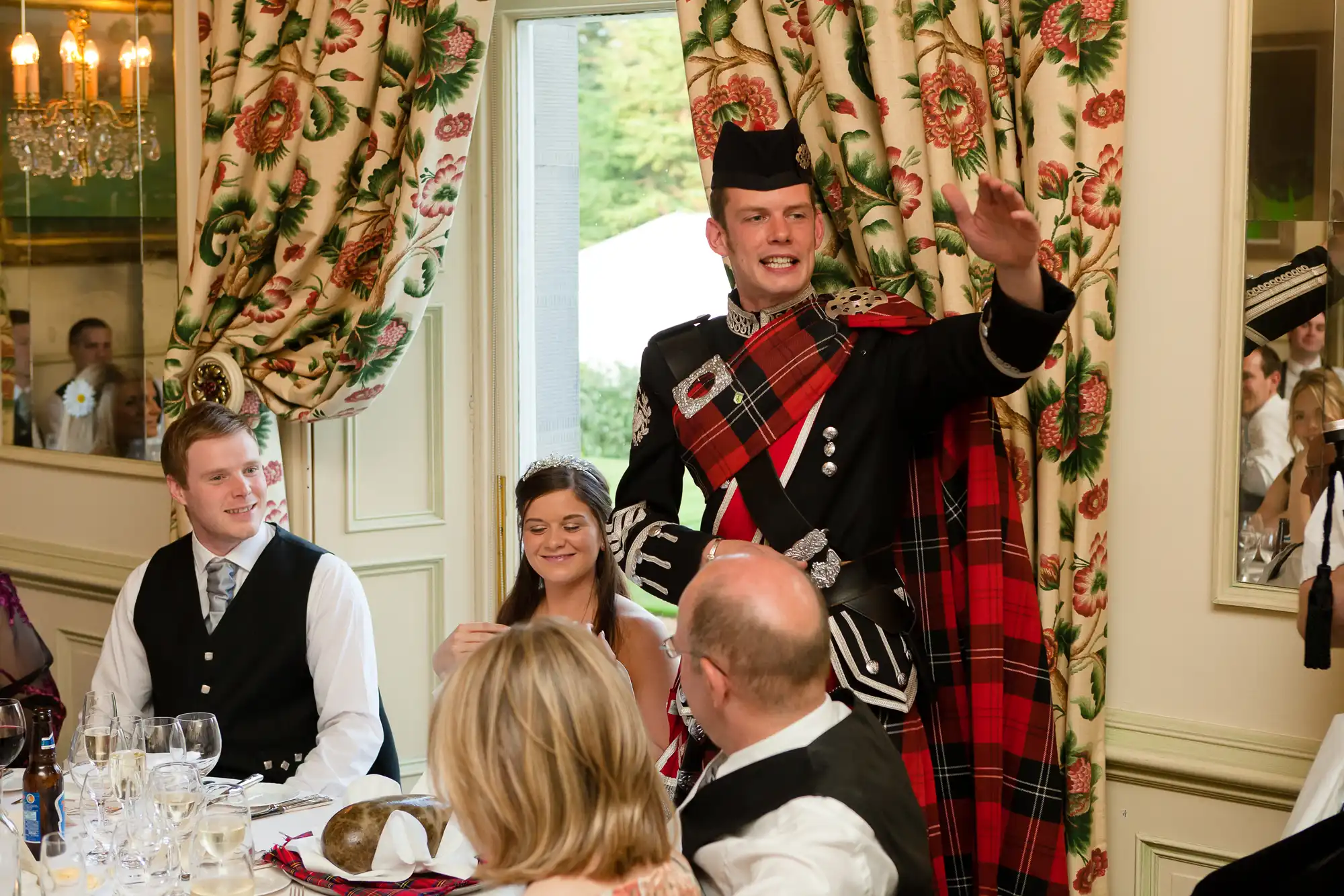 A man in traditional scottish attire addressing guests at a formal dining event, with others seated around him listening.