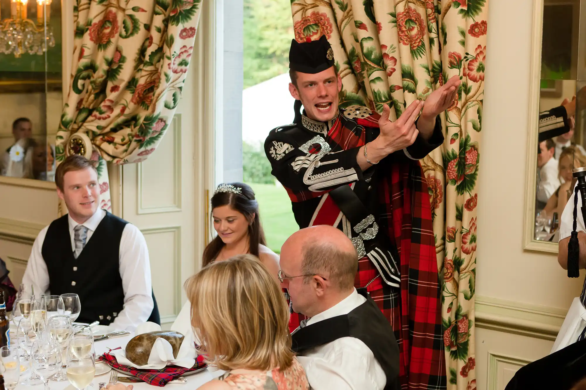 A man in traditional scottish attire presenting a haggis at a formal dining event, with guests watching in the background.