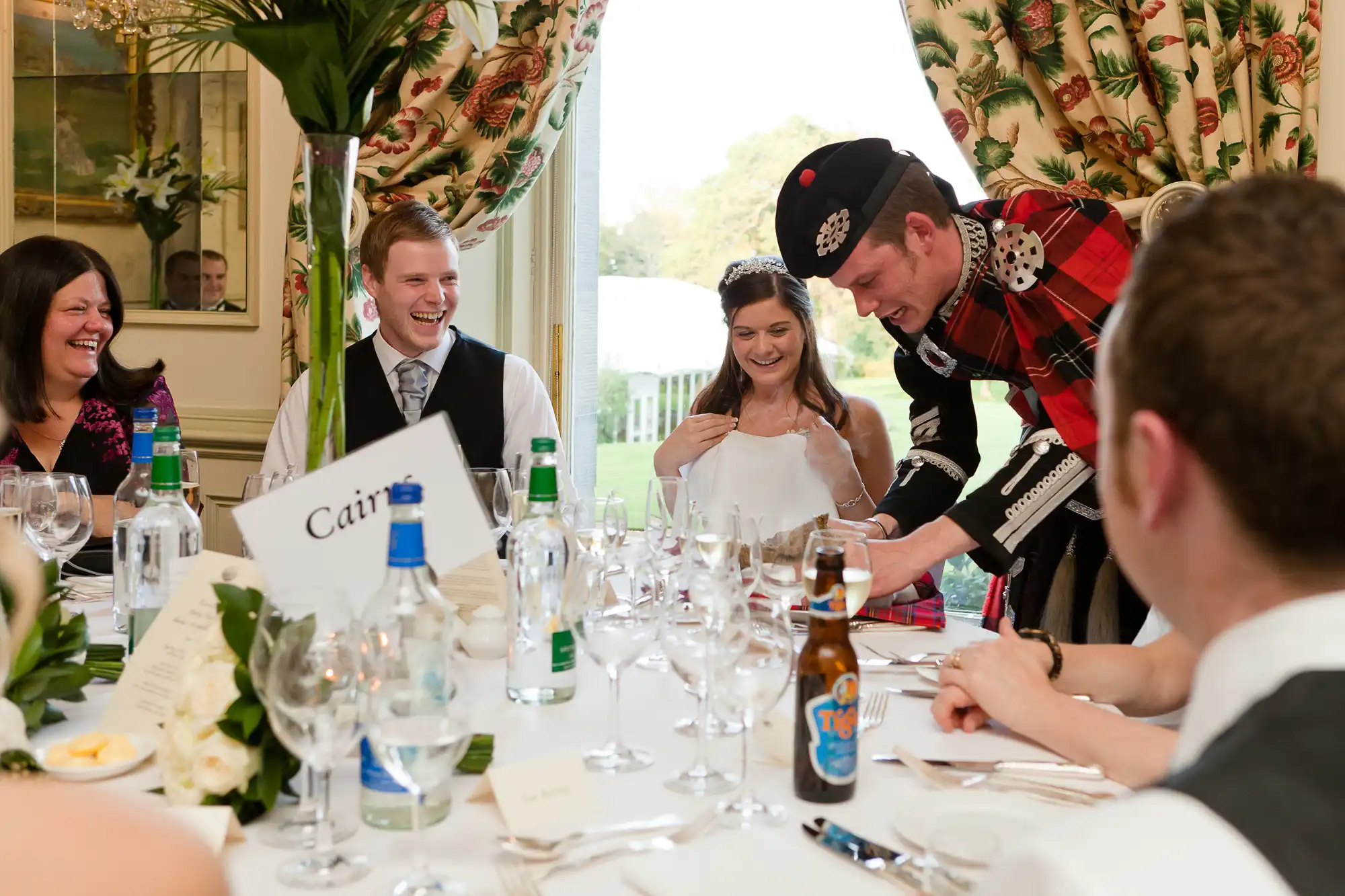 A man in traditional scottish attire interacts with guests at a wedding reception, causing laughter around a dining table.