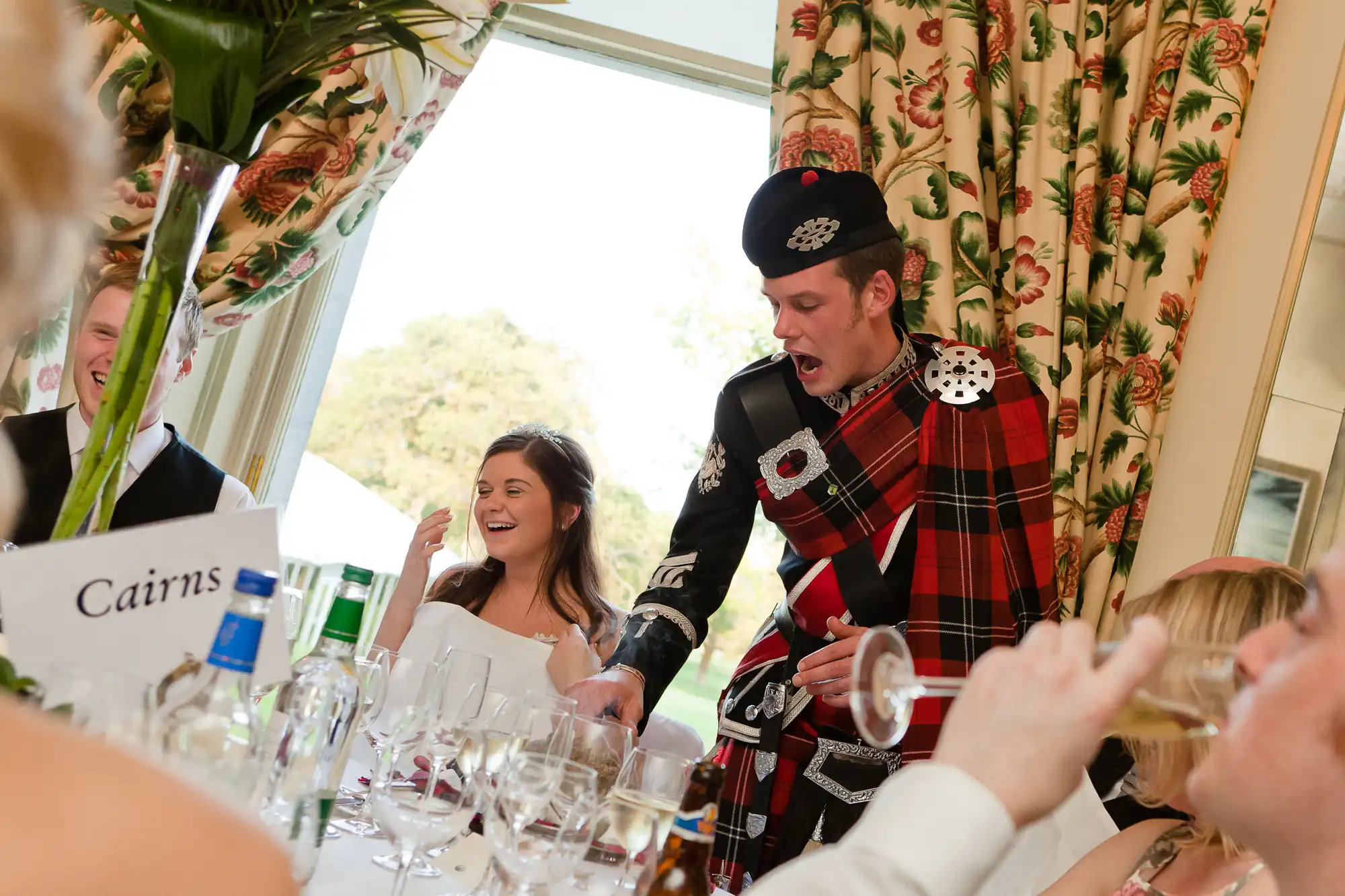 A man in scottish highland dress, including a kilt, animatedly speaks at a wedding reception, entertaining guests who are laughing and drinking.