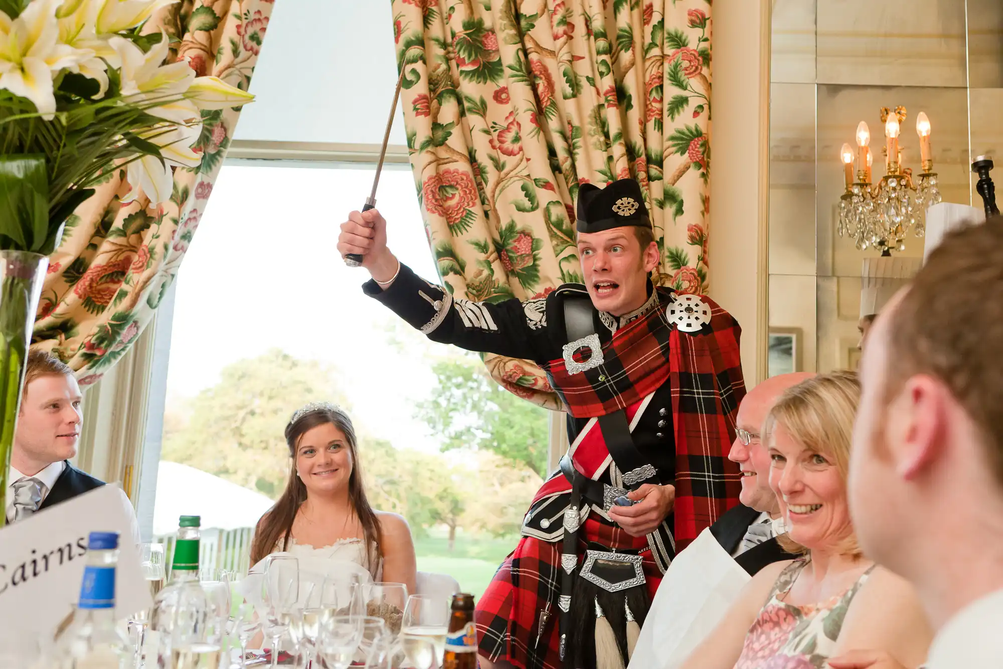 A man in a kilt and sporran enthusiastically raises a knife while giving a speech at a wedding reception, with guests smiling around him.
