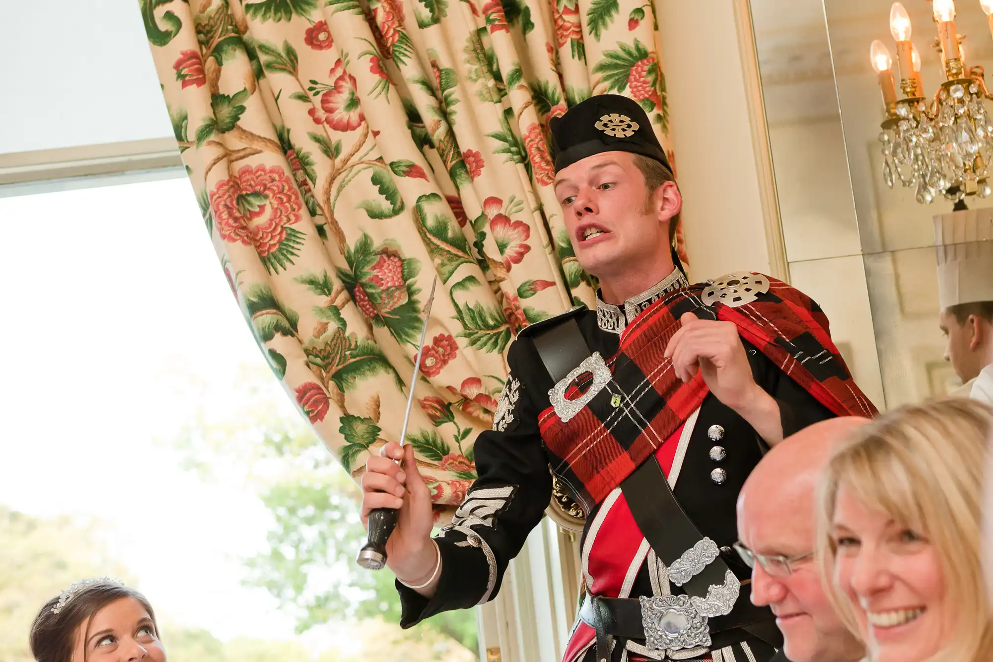 Man in traditional scottish attire playing bagpipes indoors at an event, surrounded by seated guests.
