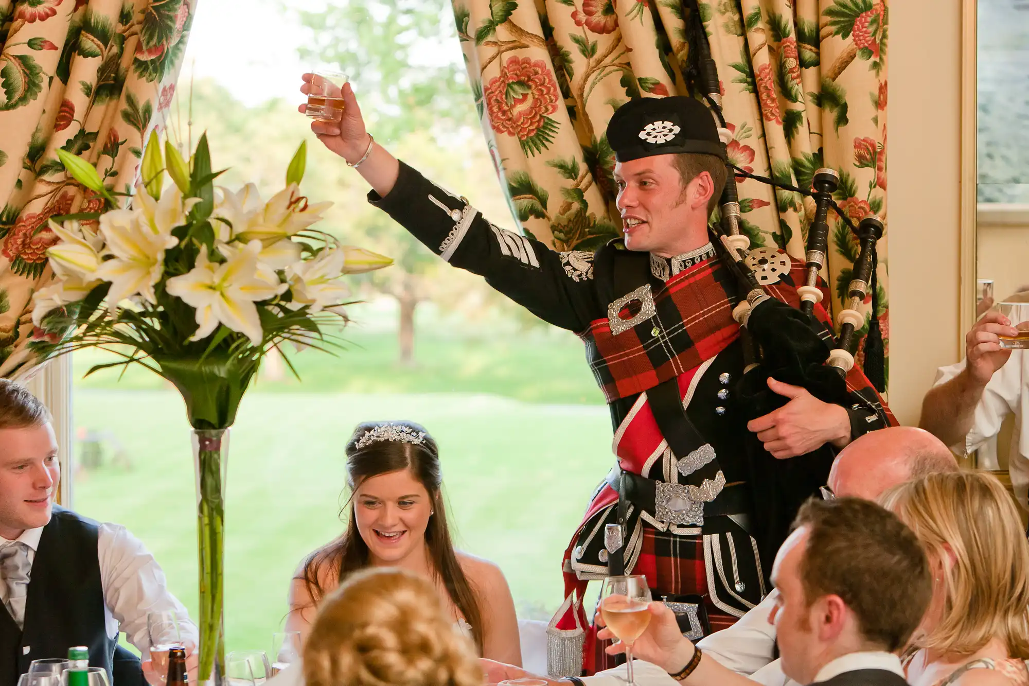A man in traditional scottish attire, holding a bagpipe, toasts at a wedding reception while guests look on.