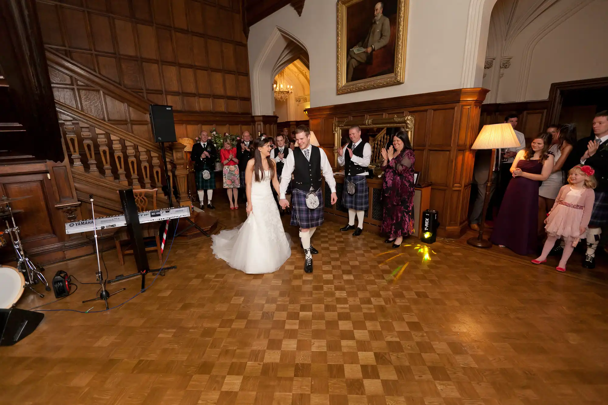 A bride and groom, the latter in a kilt, hold hands and dance in a hall with guests watching and musical instruments nearby.