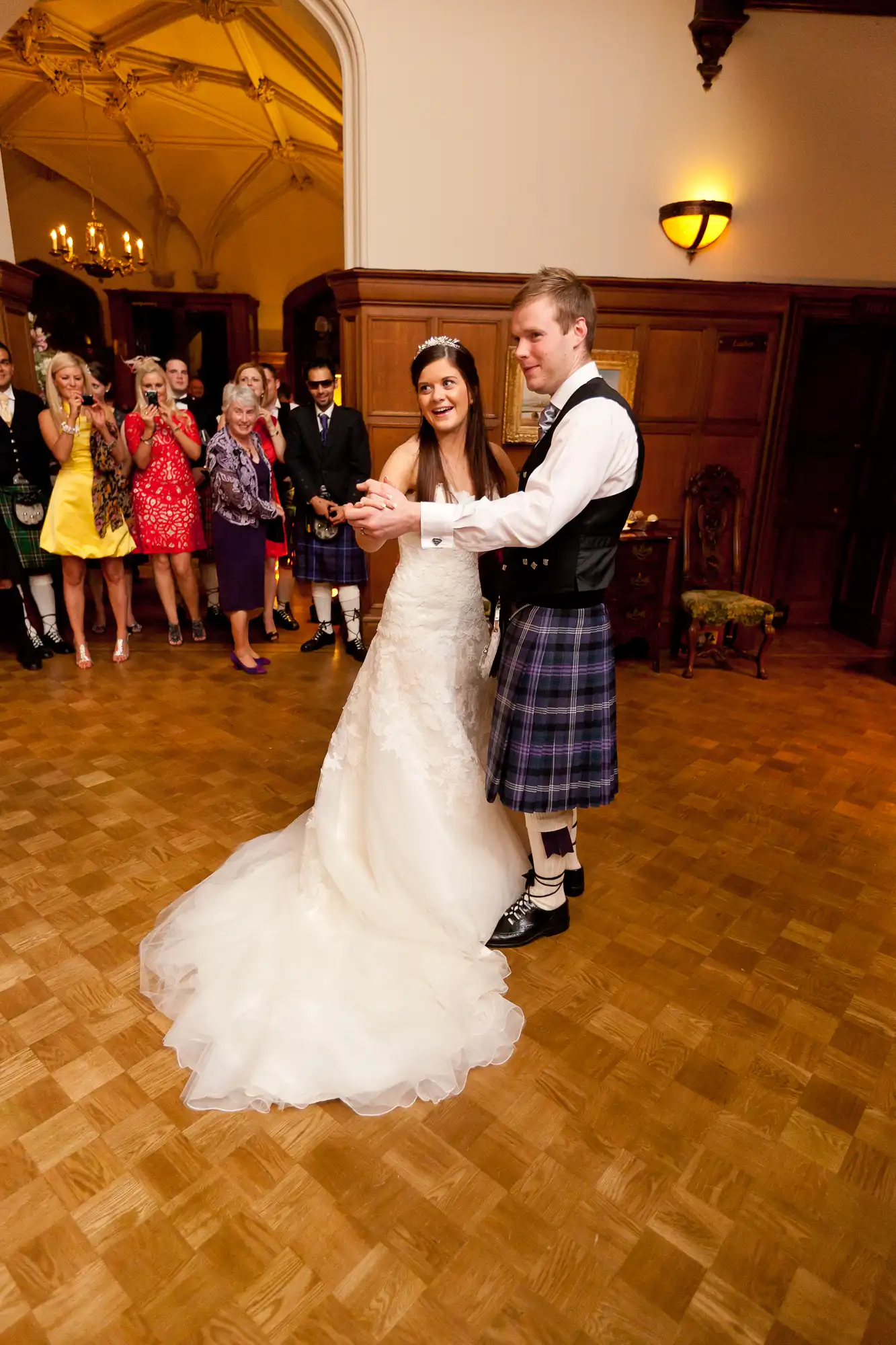 A bride in a white gown and a groom in a kilt dancing at a wedding reception, with guests watching in the background.