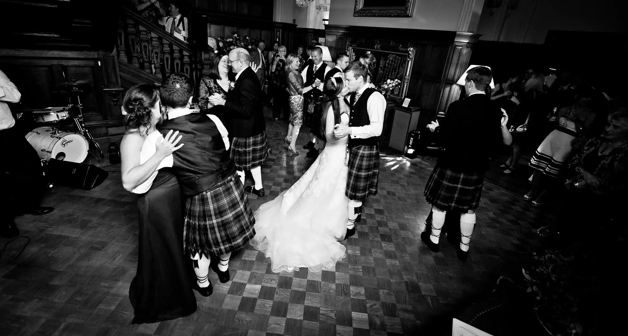 A black and white photograph of a wedding reception with guests wearing kilts dancing in a hall.
