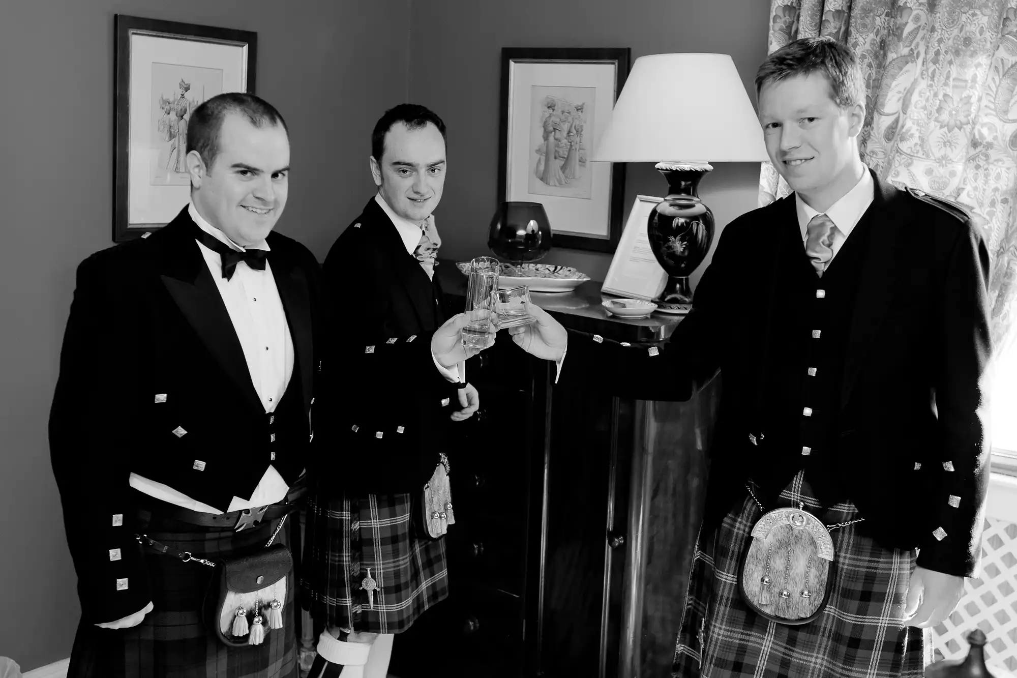 Three men in formal attire with kilts holding drinks, standing in a room with elegant decor.