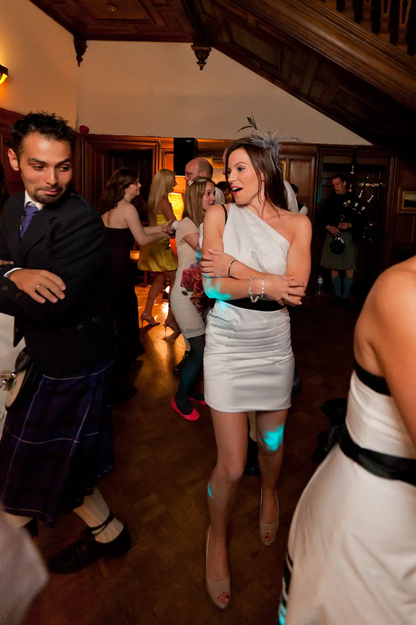 A woman in a white dress dancing at a party, surrounded by other guests, including a man in a kilt to her left.