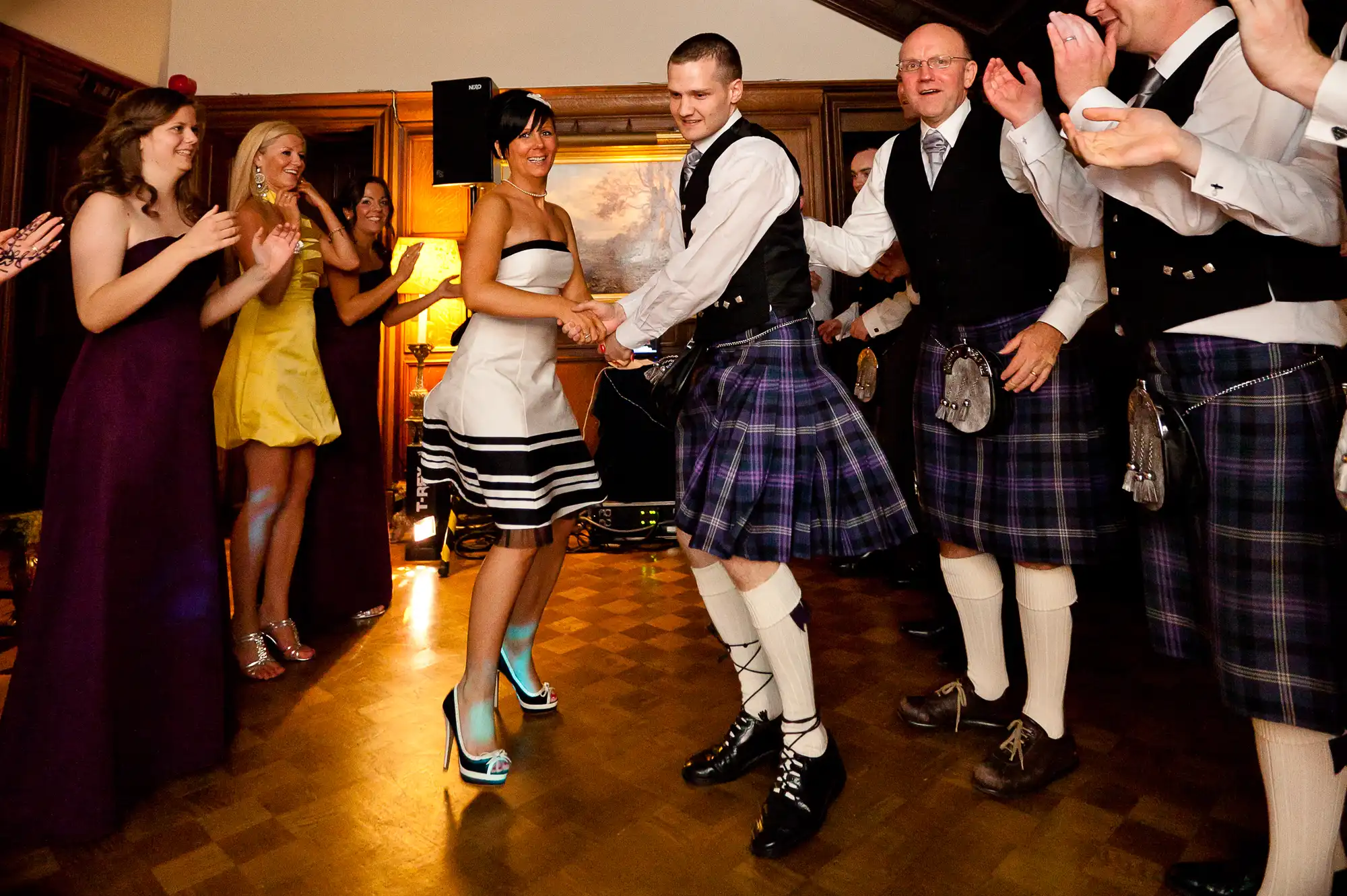 A lively dance at a scottish event with men in kilts and women in dresses, guests clapping joyfully around them.