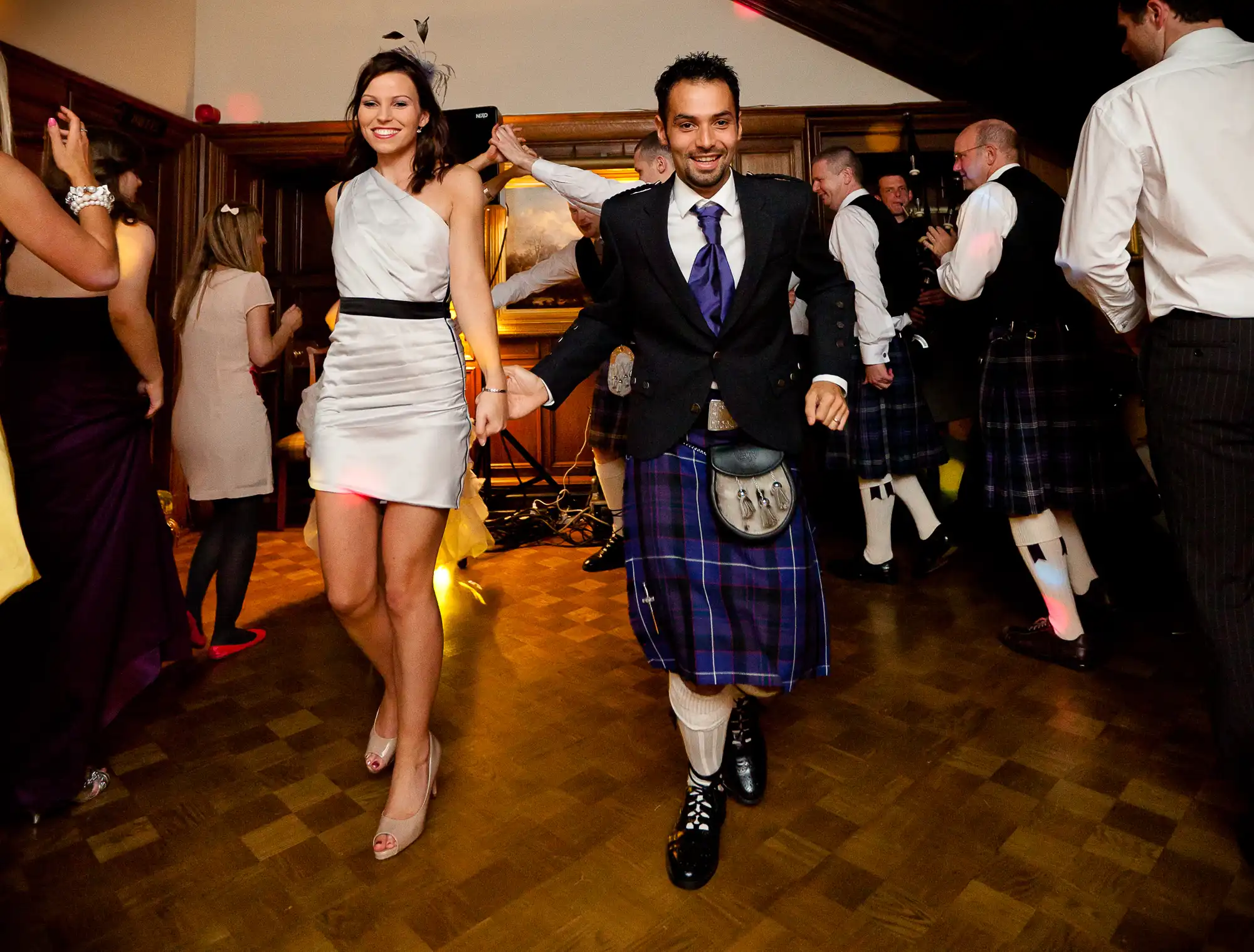 A couple dancing joyfully at a wedding reception, the man in a traditional kilt and the woman in a white dress, surrounded by other guests in kilts.