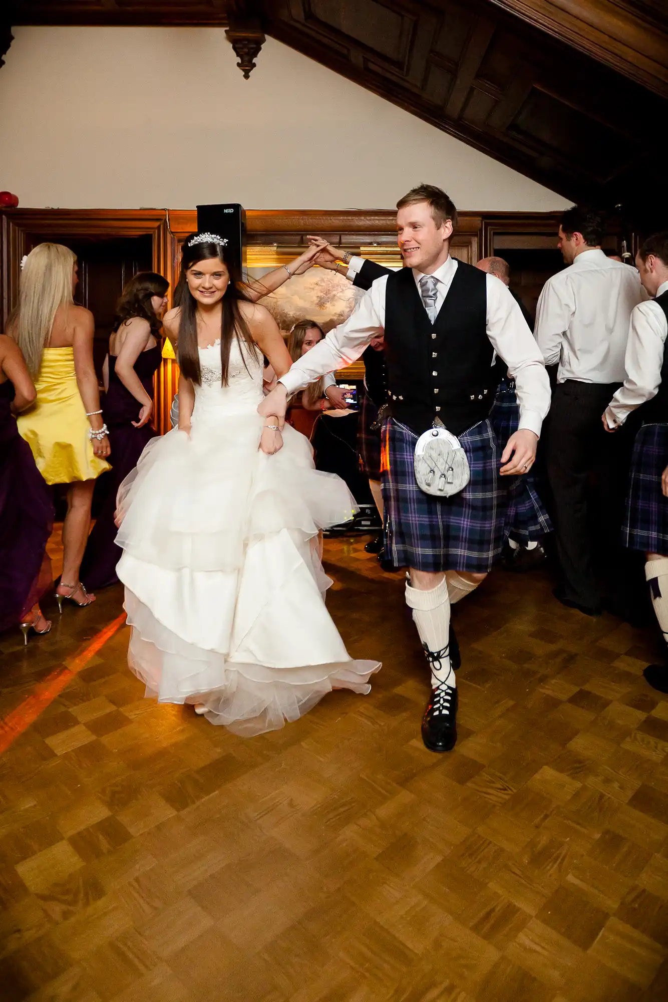 A bride in a white gown and a groom in a traditional scottish kilt and jacket dance joyfully at a wedding reception.