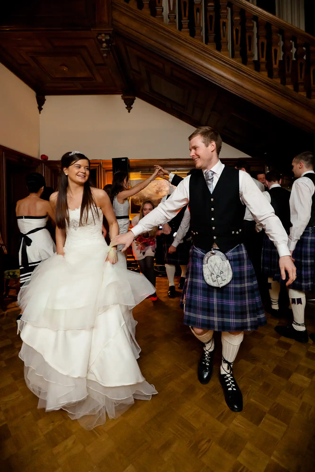 A bride in a white gown and a groom in a kilt and sporran holding hands and smiling during a dance at a wedding reception with guests in the background.