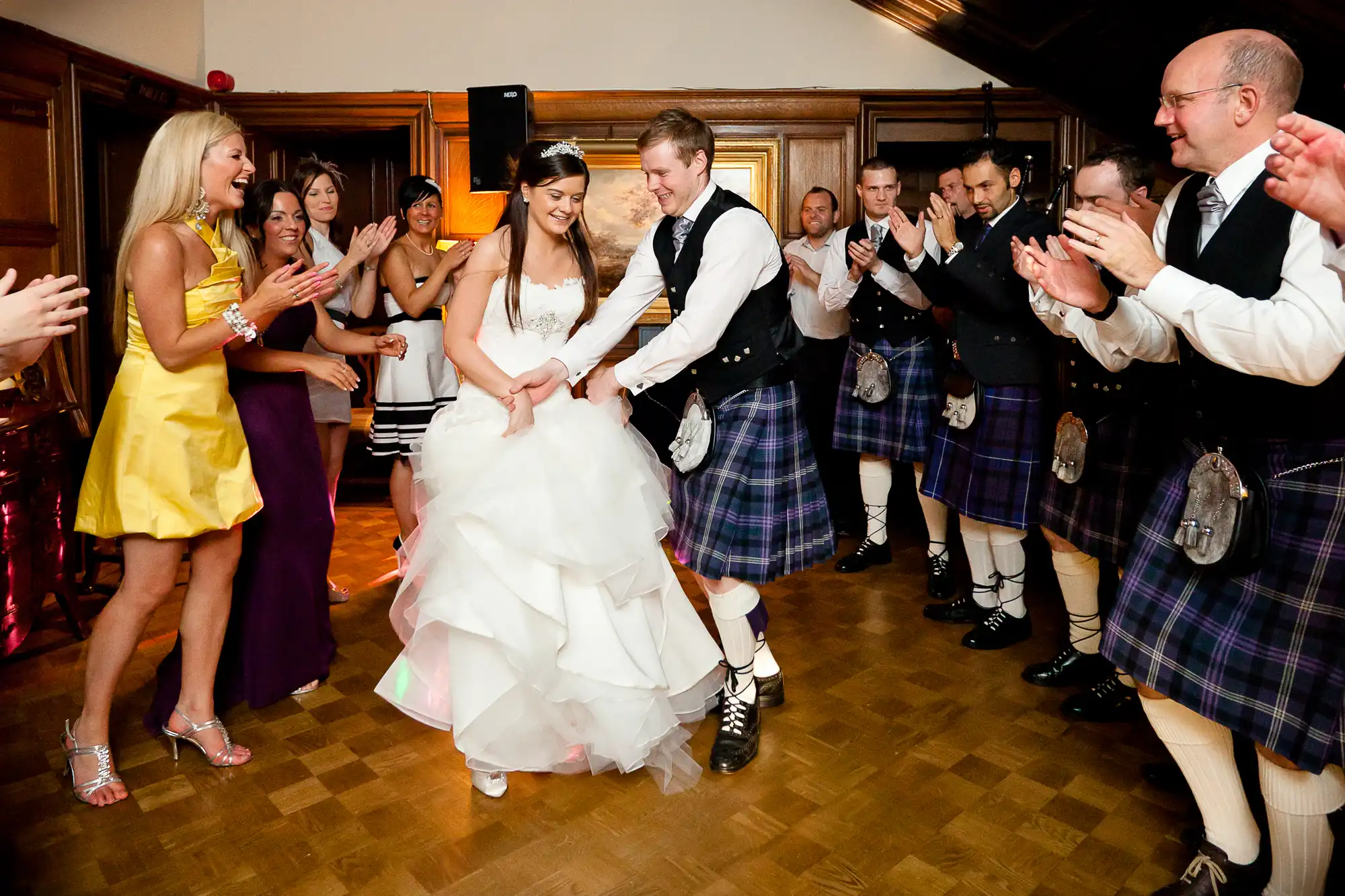 Bride and groom dancing in a hall, surrounded by clapping guests in formal wear and kilts, expressing joy during a wedding reception.