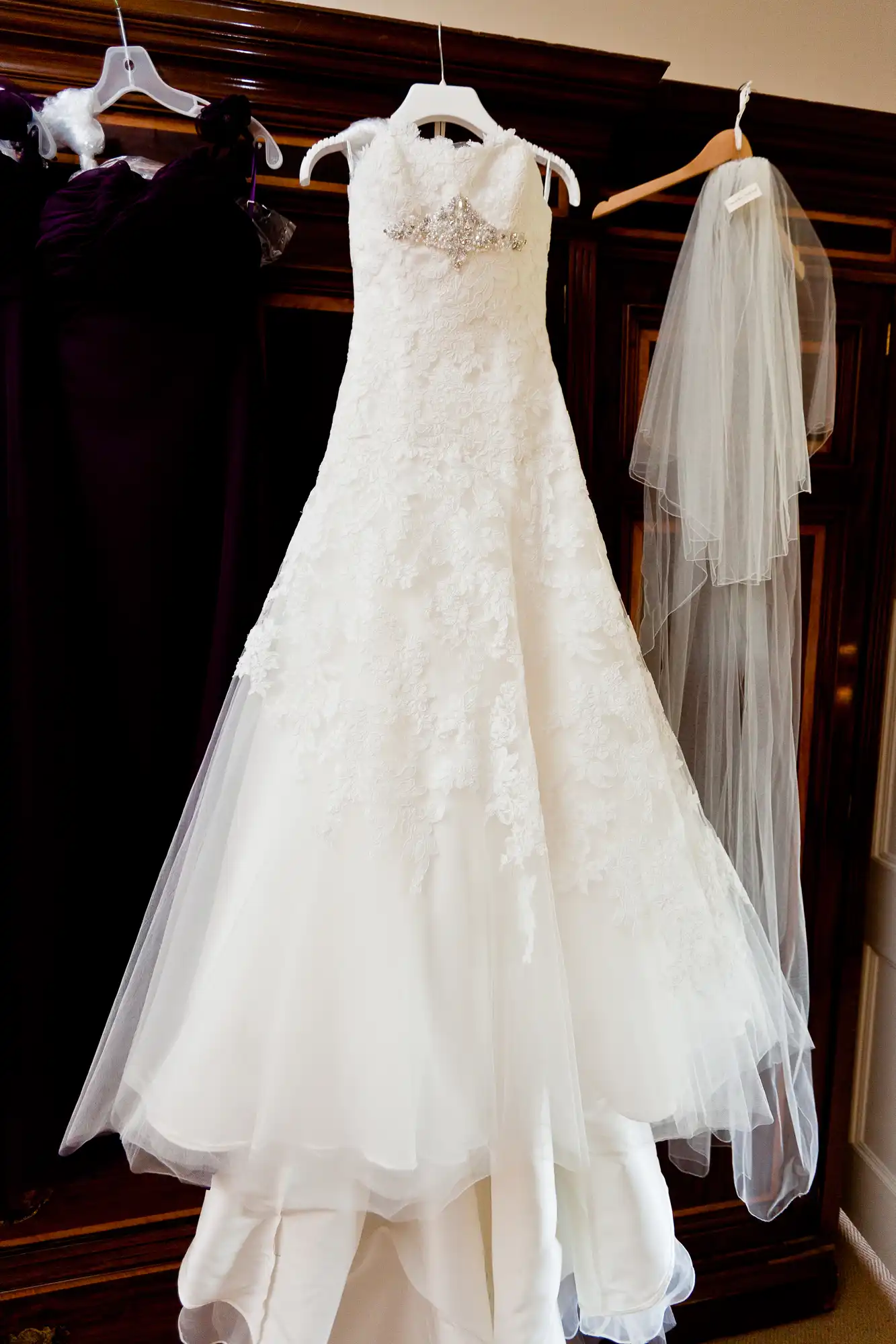 A white wedding dress with lace details and a beaded bodice hangs alongside a veil on wooden hangers against a dark background.