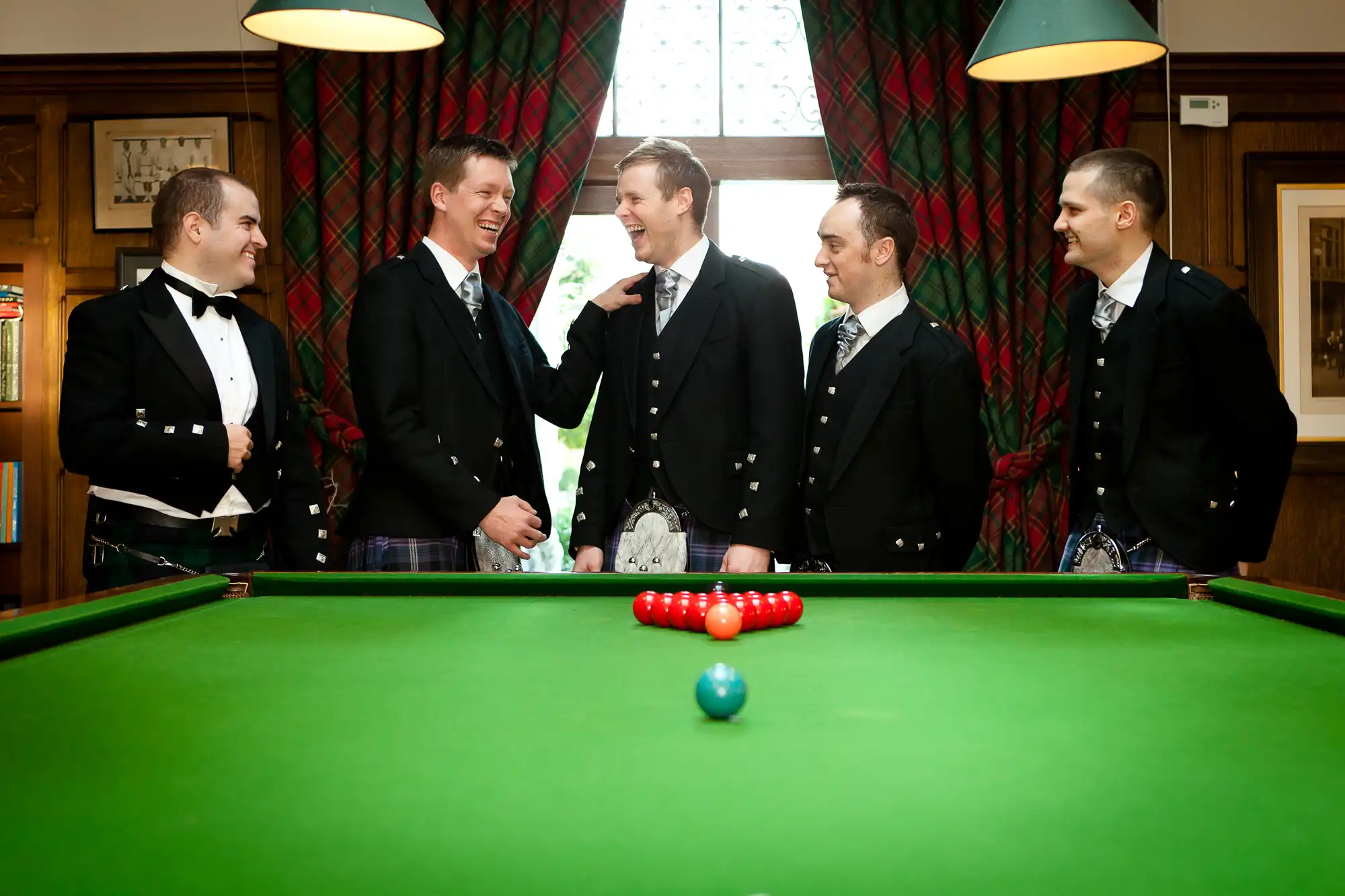 Five men in kilts and tuxedo jackets smiling and chatting around a billiards table in a warmly lit room.
