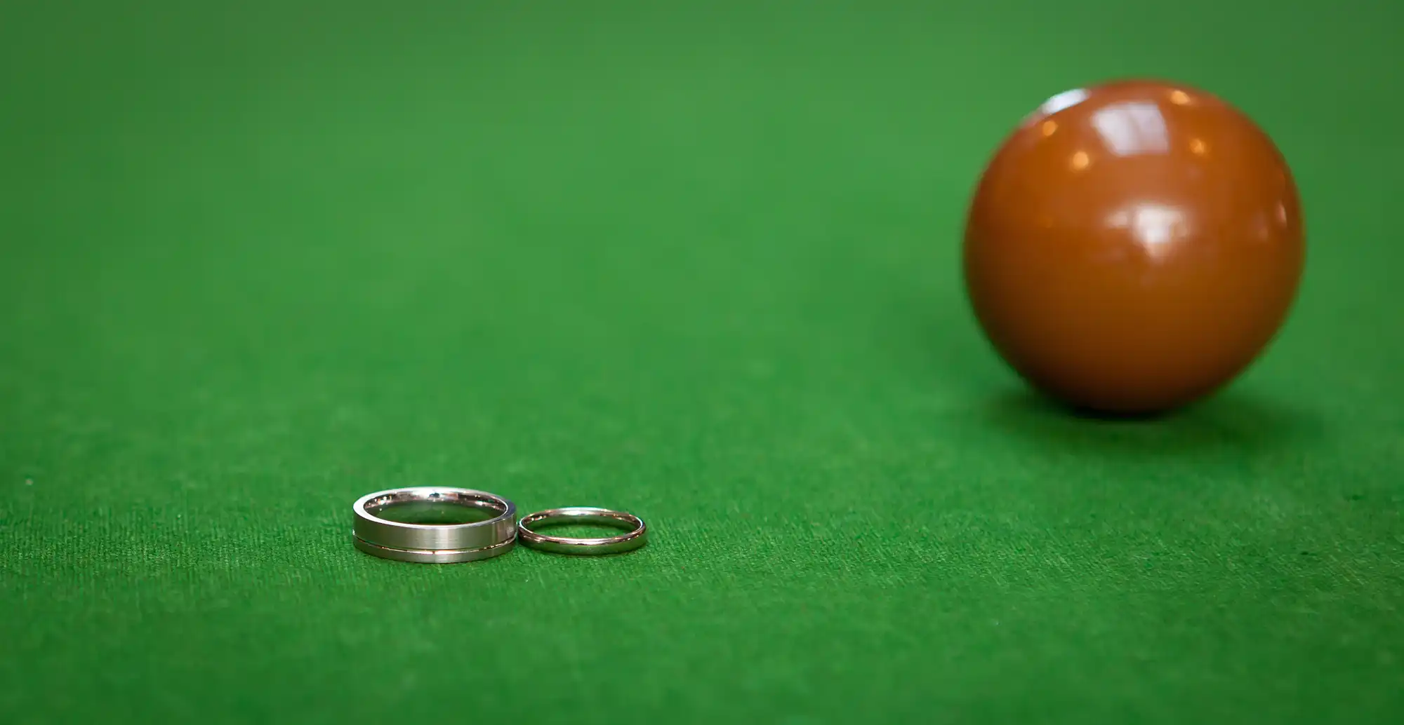 Two wedding rings placed on a green surface near a shiny orange billiard ball.