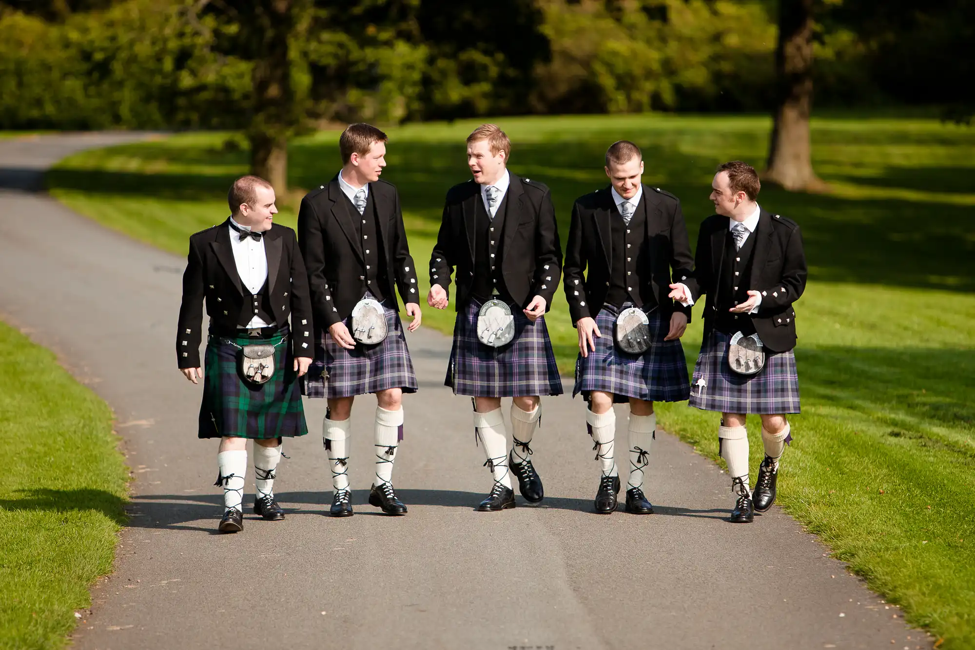 Five men in traditional scottish kilts and jackets walking along a park path, chatting and smiling on a sunny day.