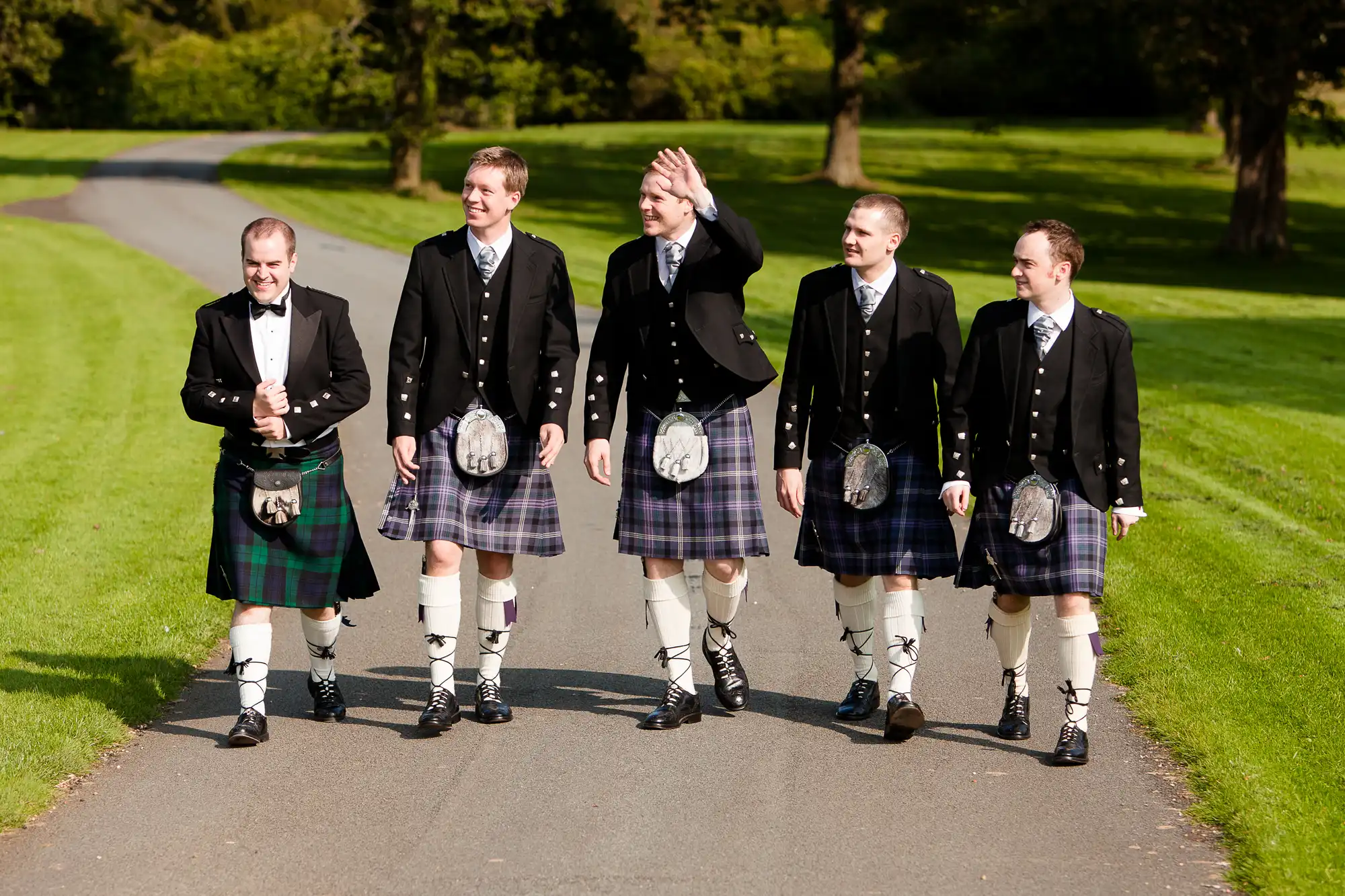 Five men in traditional scottish attire, including kilts and sporran, walking along a sunlit park path, smiling and gesturing.