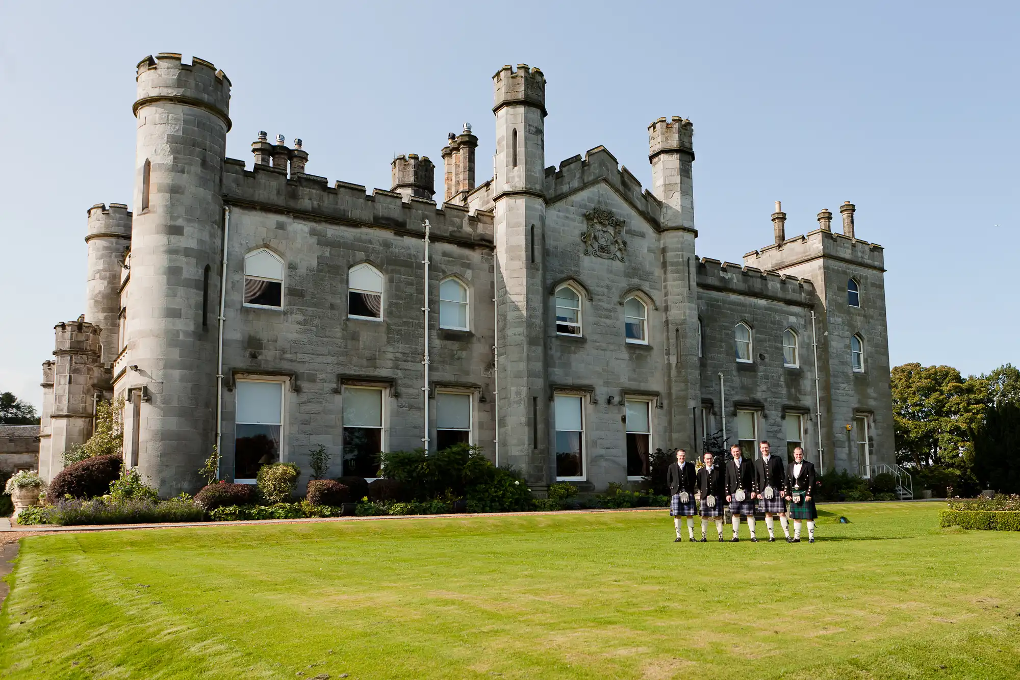 A row of seven people in traditional scottish attire standing in front of an historic stone castle with turrets under a clear sky.