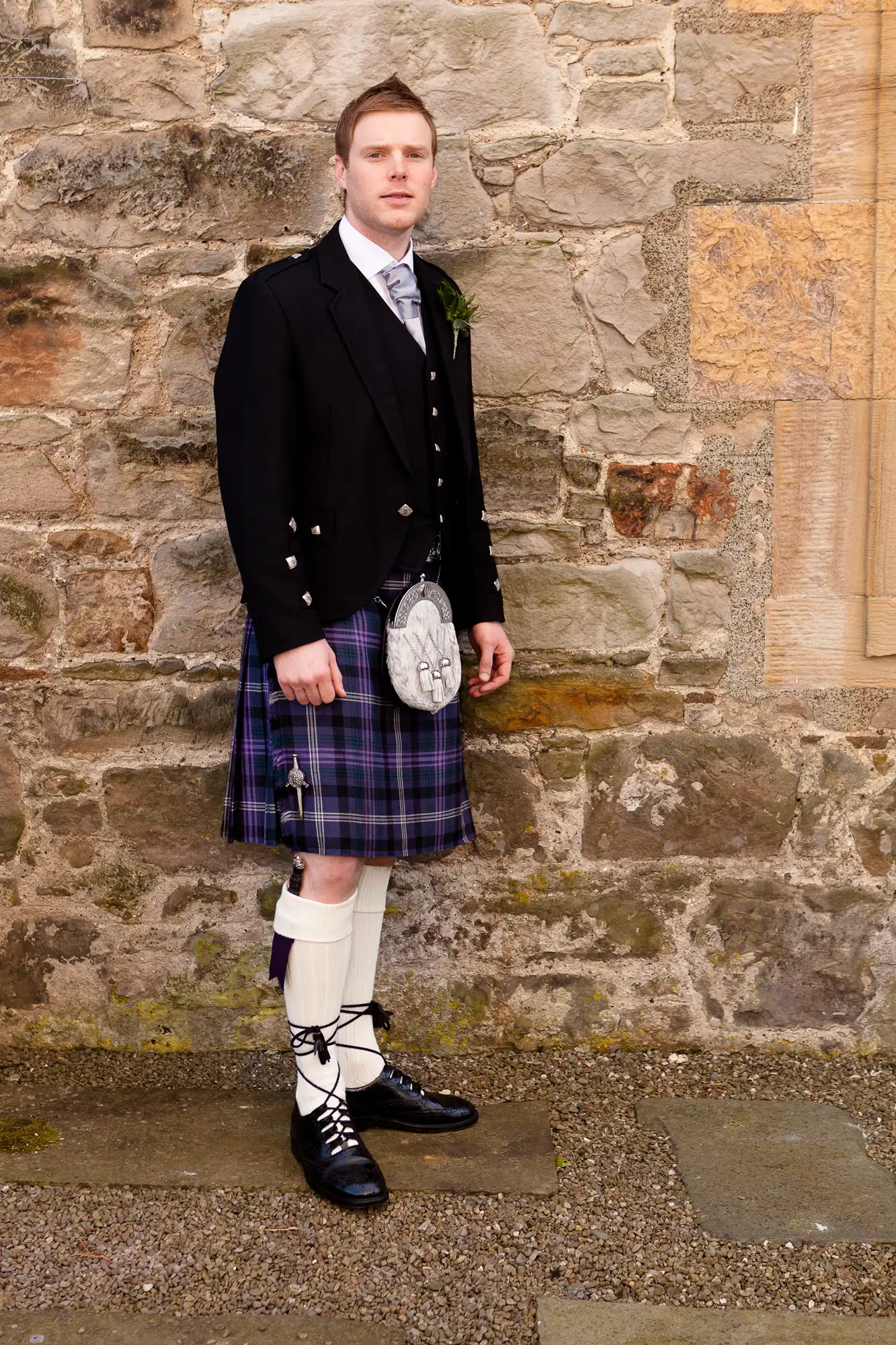 A man in traditional scottish attire, including a kilt, sporran, and black jacket, stands against a stone wall.