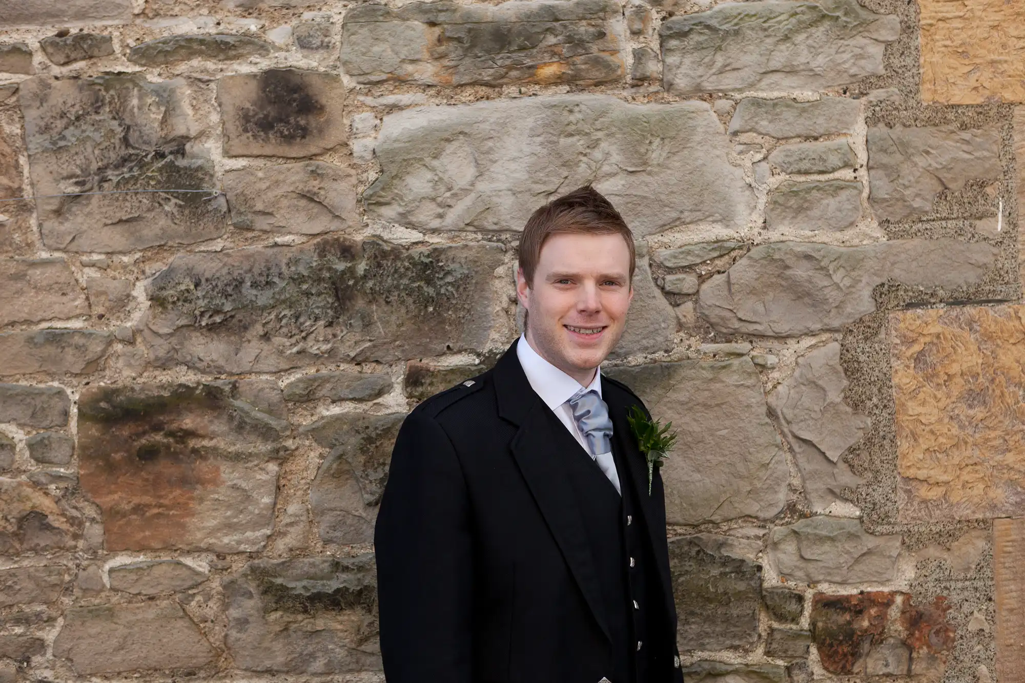 A young man in a black suit and tie smiling in front of a textured stone wall.