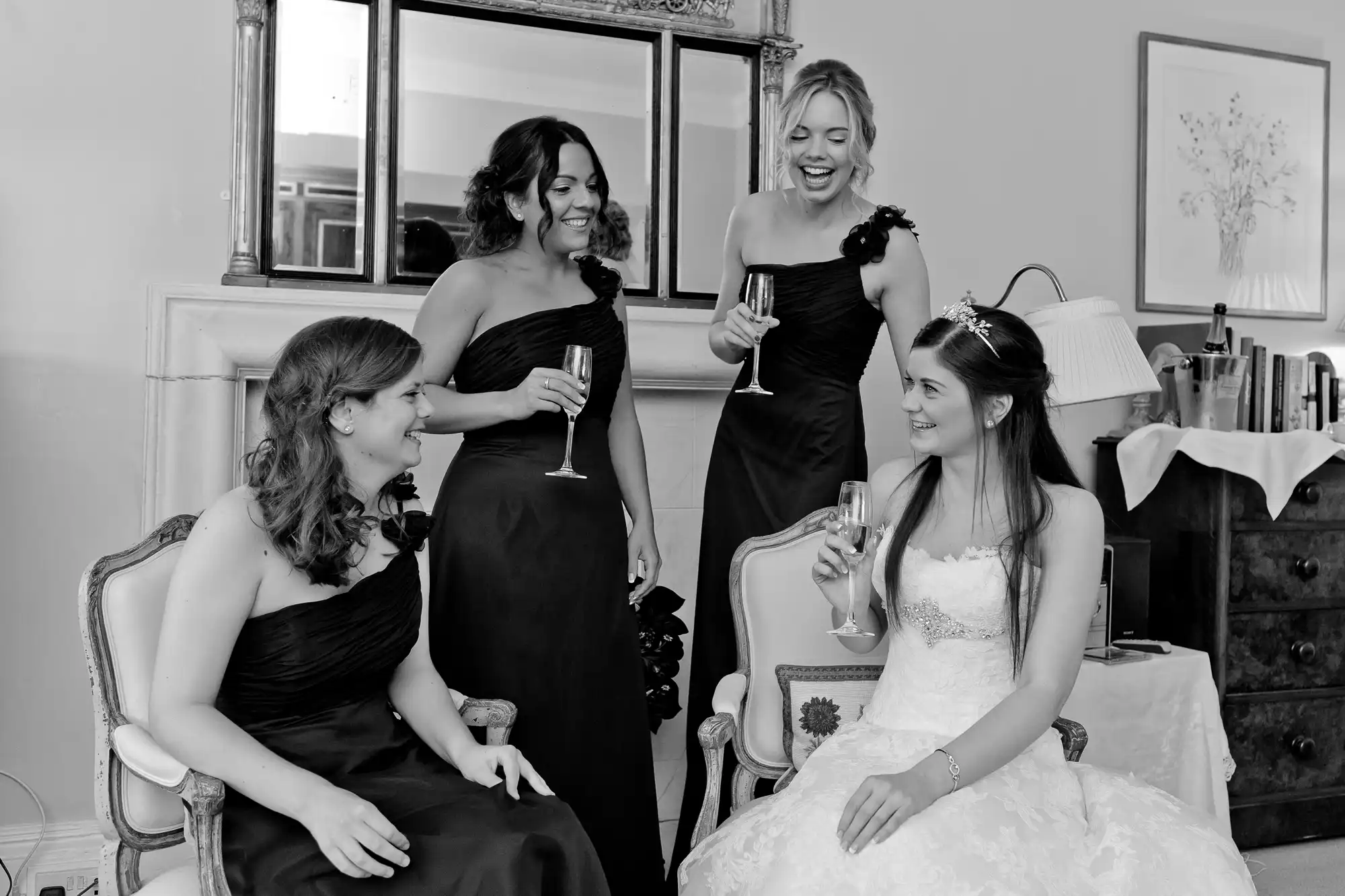 Four women in elegant dresses, one in a bridal gown, cheerfully chatting and holding champagne glasses in a room with a vintage dresser.