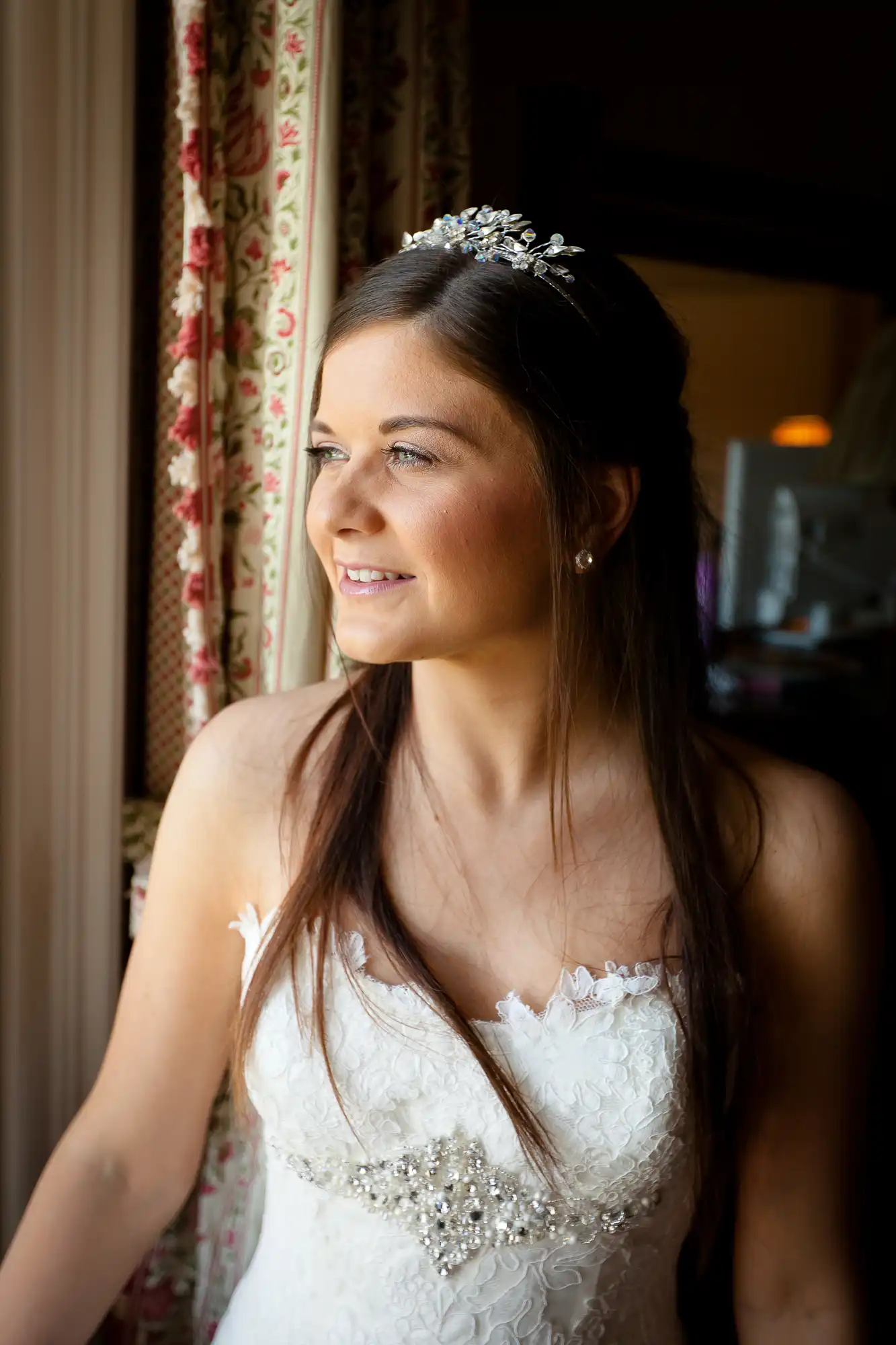 A bride in a detailed white gown and a bejeweled headpiece smiles softly, looking out a window.