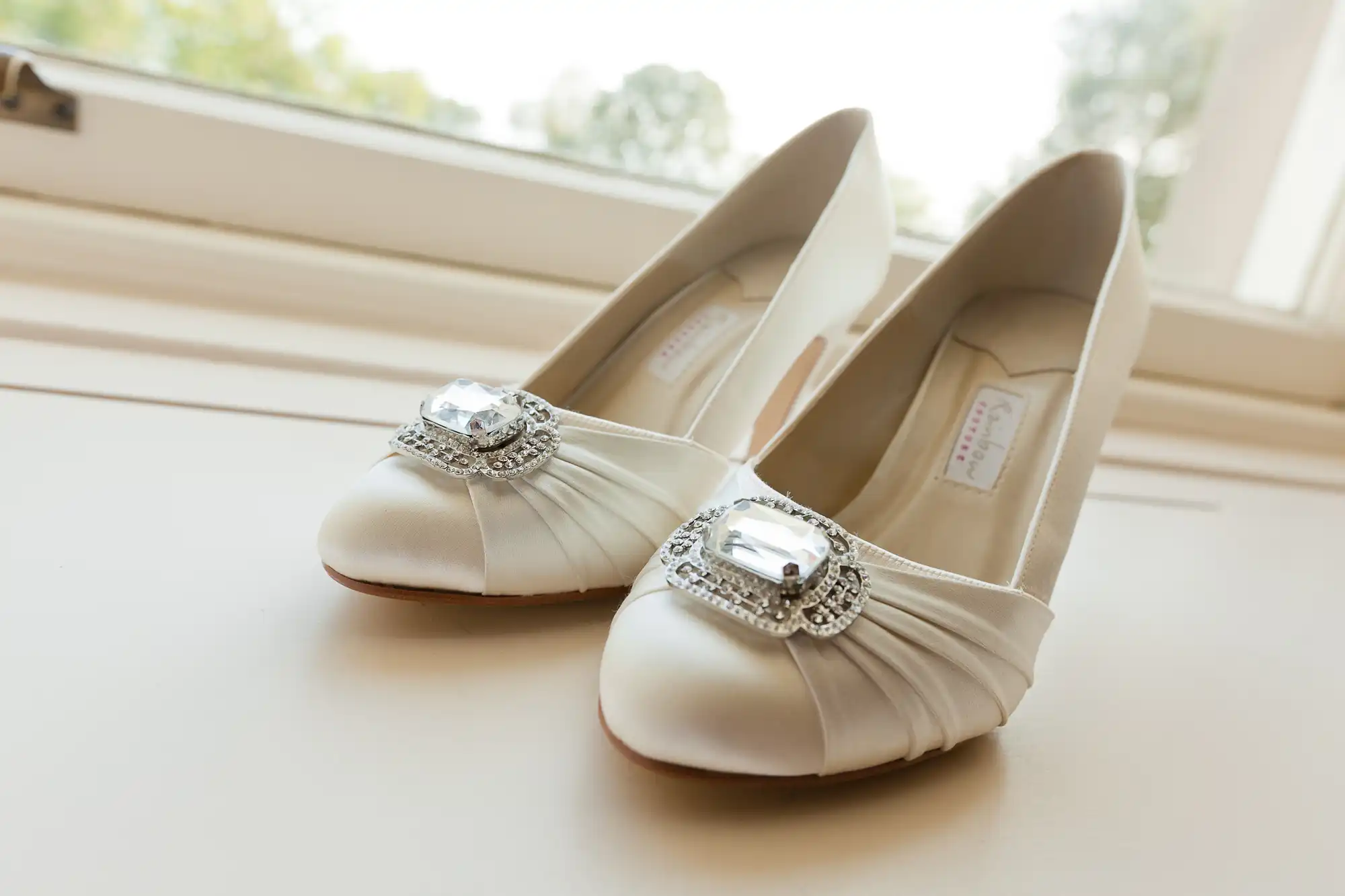 A pair of elegant white bridal shoes with rhinestone embellishments, placed by a window casting soft natural light.