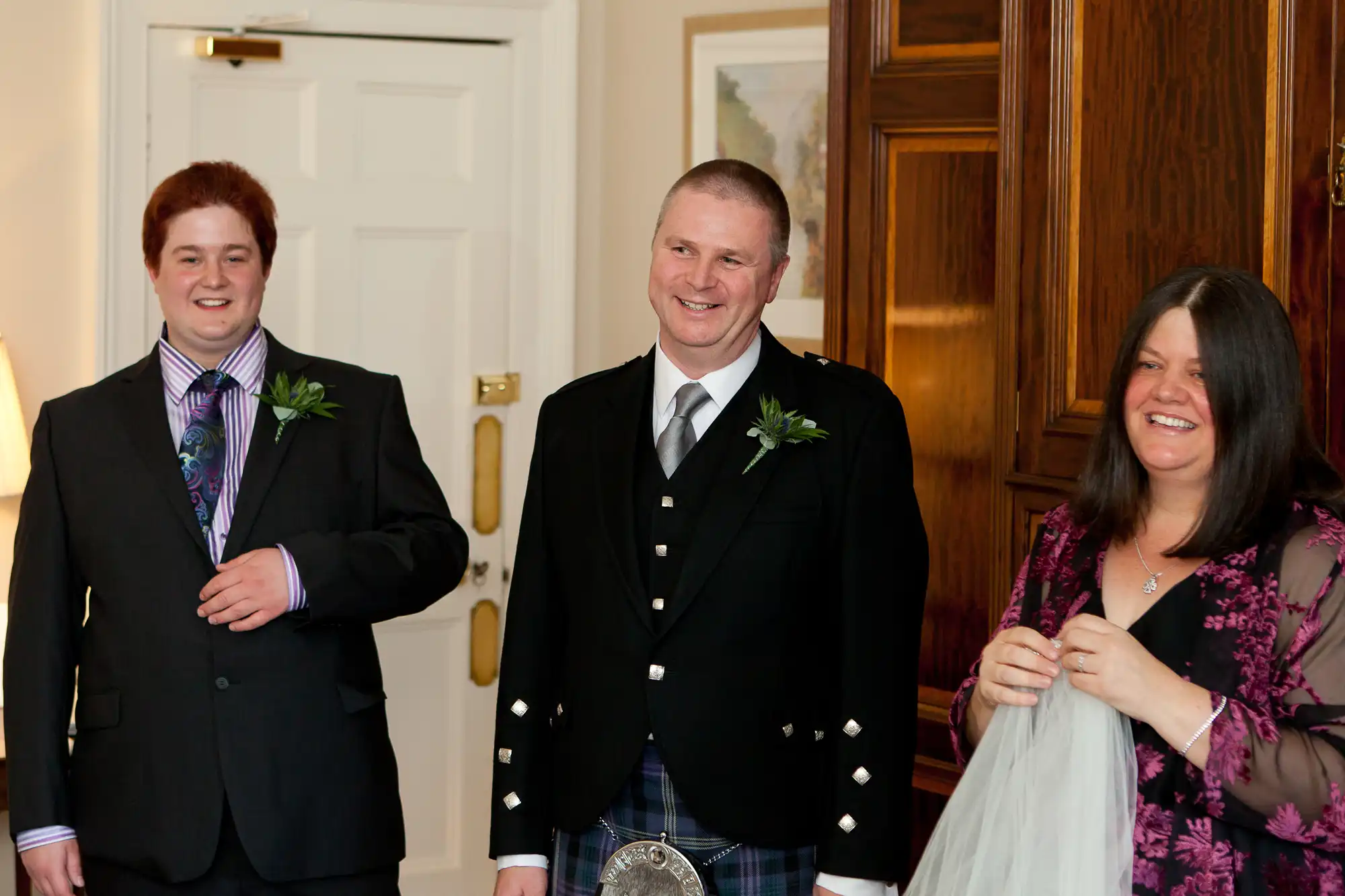 Three people smiling in a room, one in traditional scottish attire with a kilt, standing near an open door.
