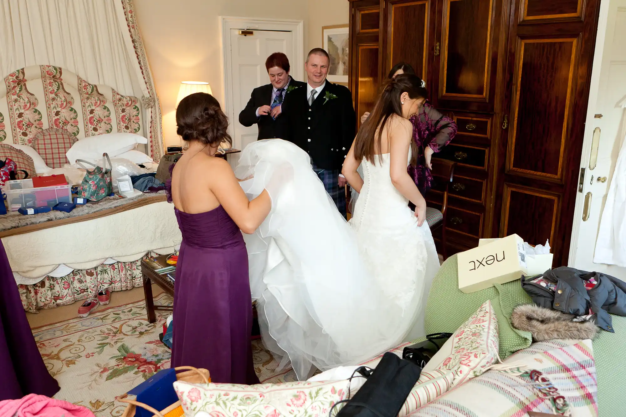 A bride in a white gown is helped by two women in a cluttered room, with a man in a kilt smiling in the background.