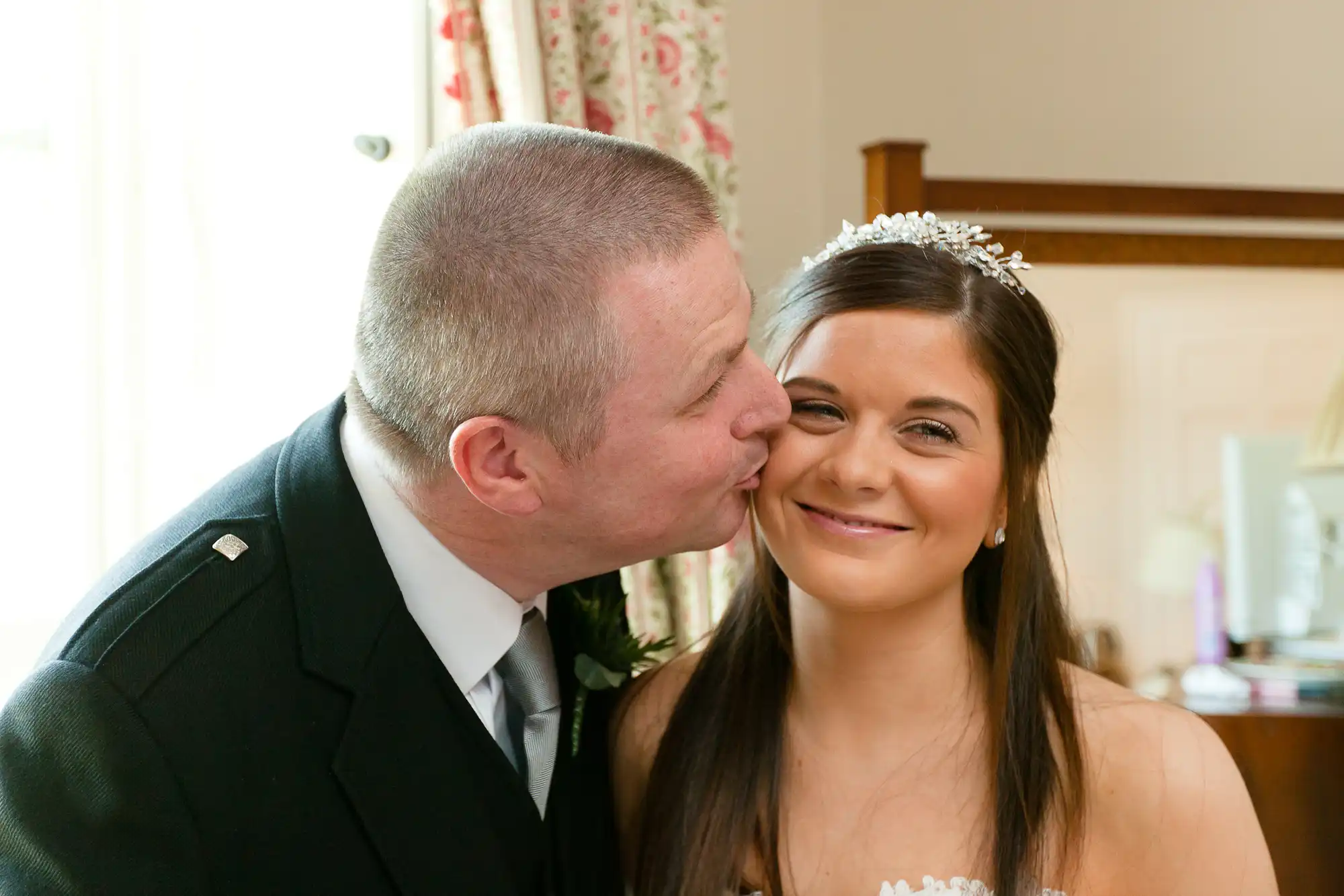 A man in a military uniform kisses a smiling bride on the cheek in a warmly lit room.