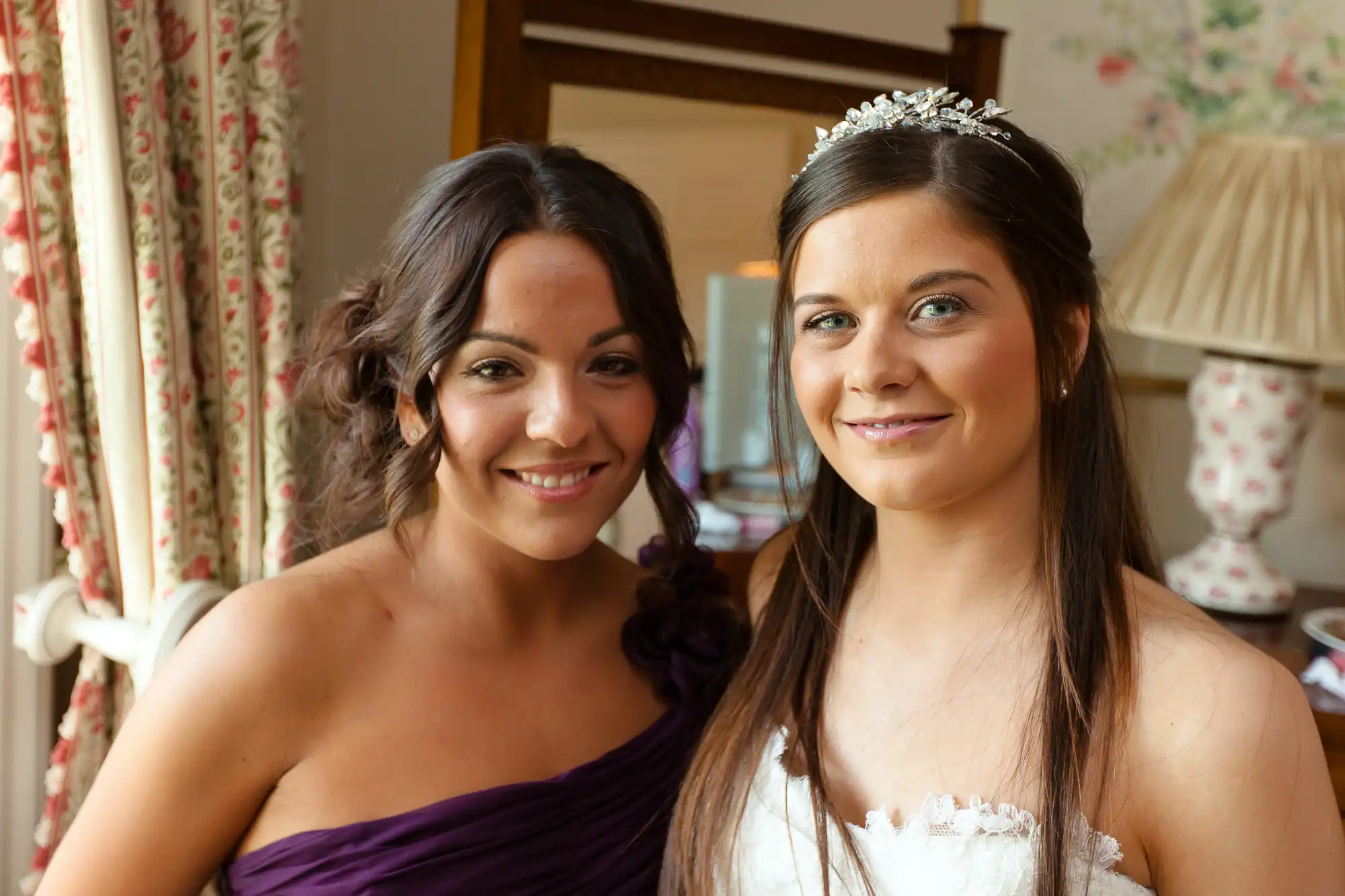 Two women smiling indoors, one in a white bridal gown with a tiara and the other in a purple dress.
